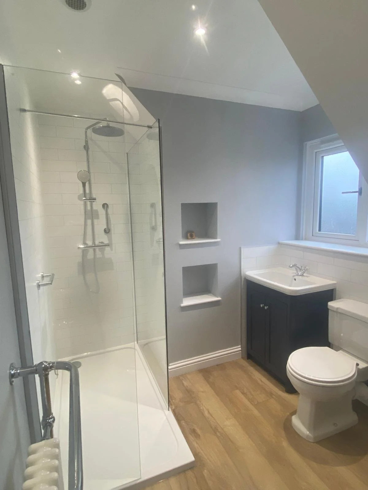 A modern bathroom with a walk-in shower, a small vanity with a sink, a toilet, and a frosted window. The floor is wood, and the walls are painted light gray.