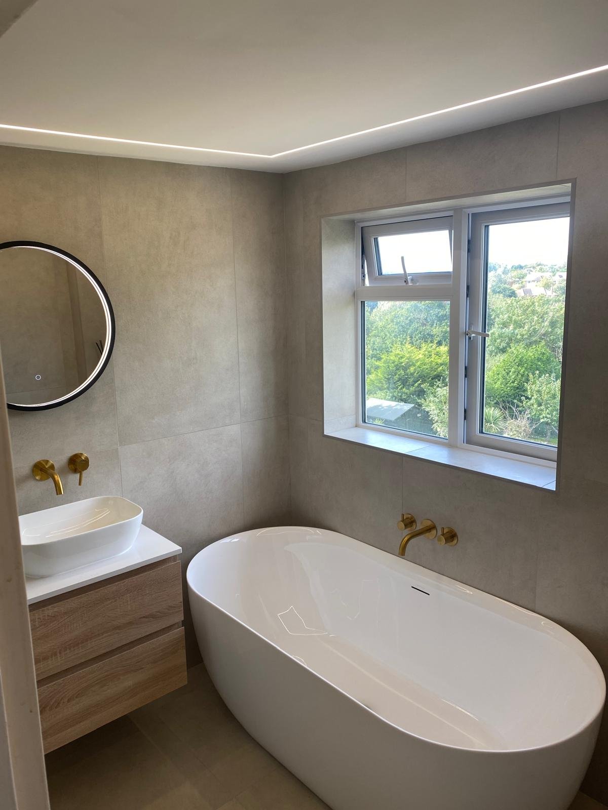 Beige tiled bathroom with standalone bath and gold taps installed on the wall. There is a black circle mirror on the wall and white sink on top of wooden drawers