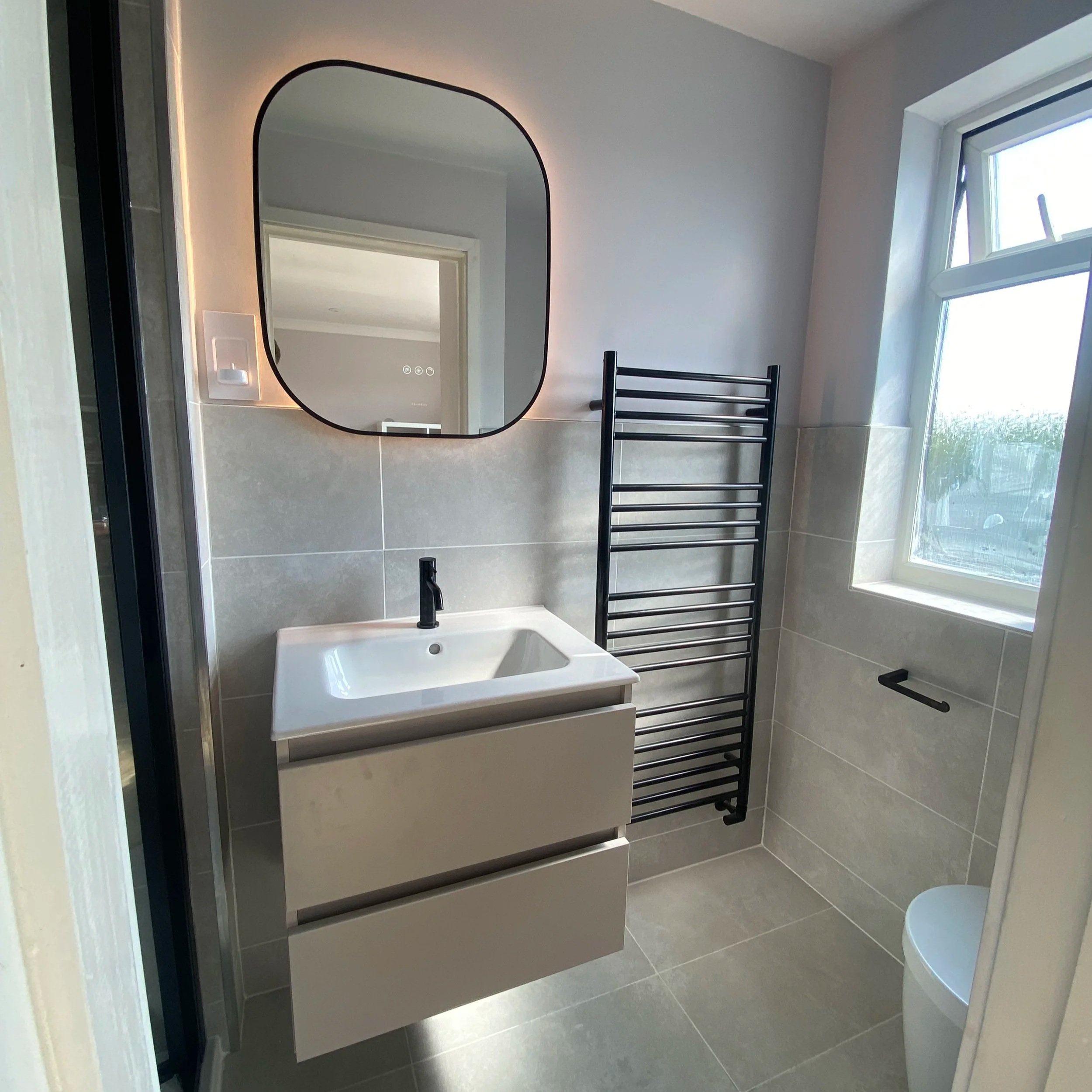 Modern bathroom with a wall-mounted sink, black faucet, and a large backlit mirror. Black heated towel rail, window with outdoor view, and grey tiled walls and floor.