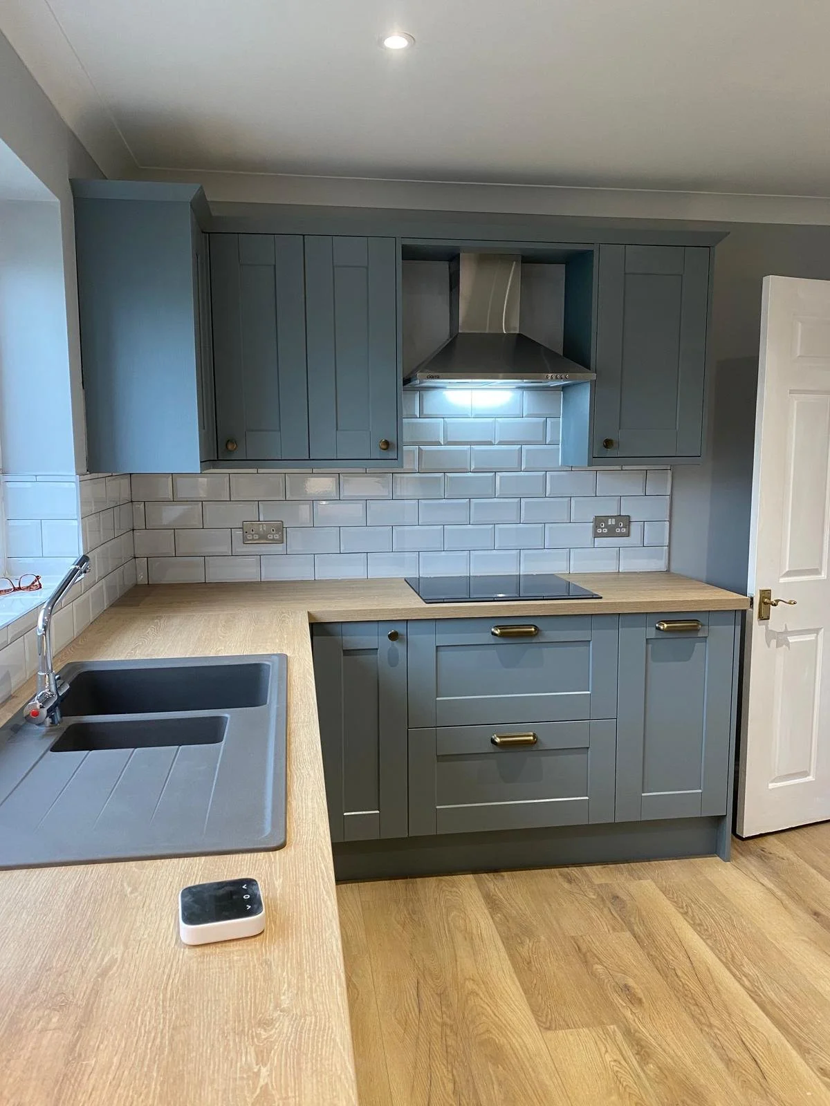 Grey shaker style kitchen cabinets with wooden work tops. There is a grey sink with silver tap, as well as silver extractor fan and white door