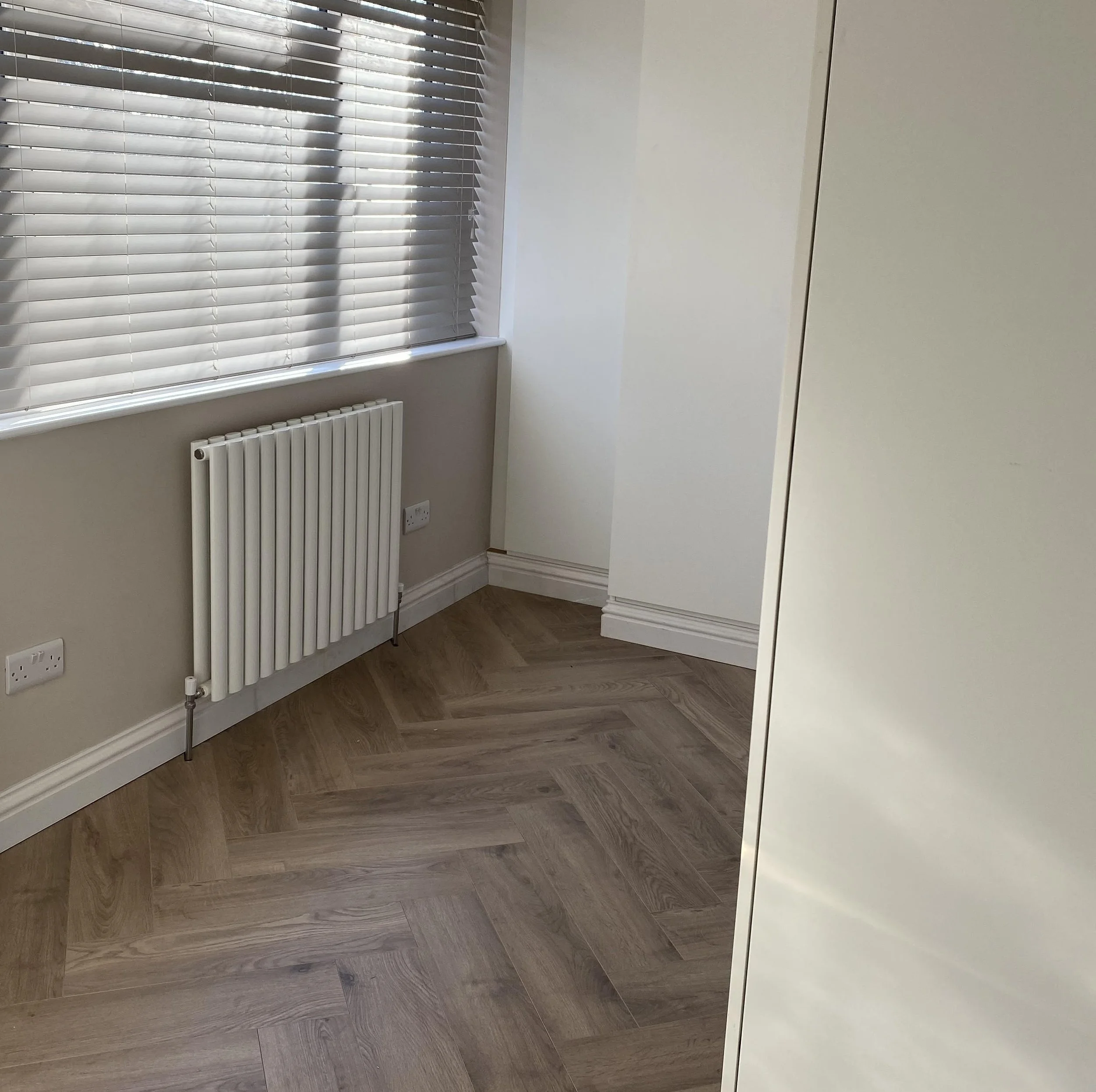 A corner of a room with a window with blinds, a white radiator, and light-colored wooden herringbone-patterned flooring.