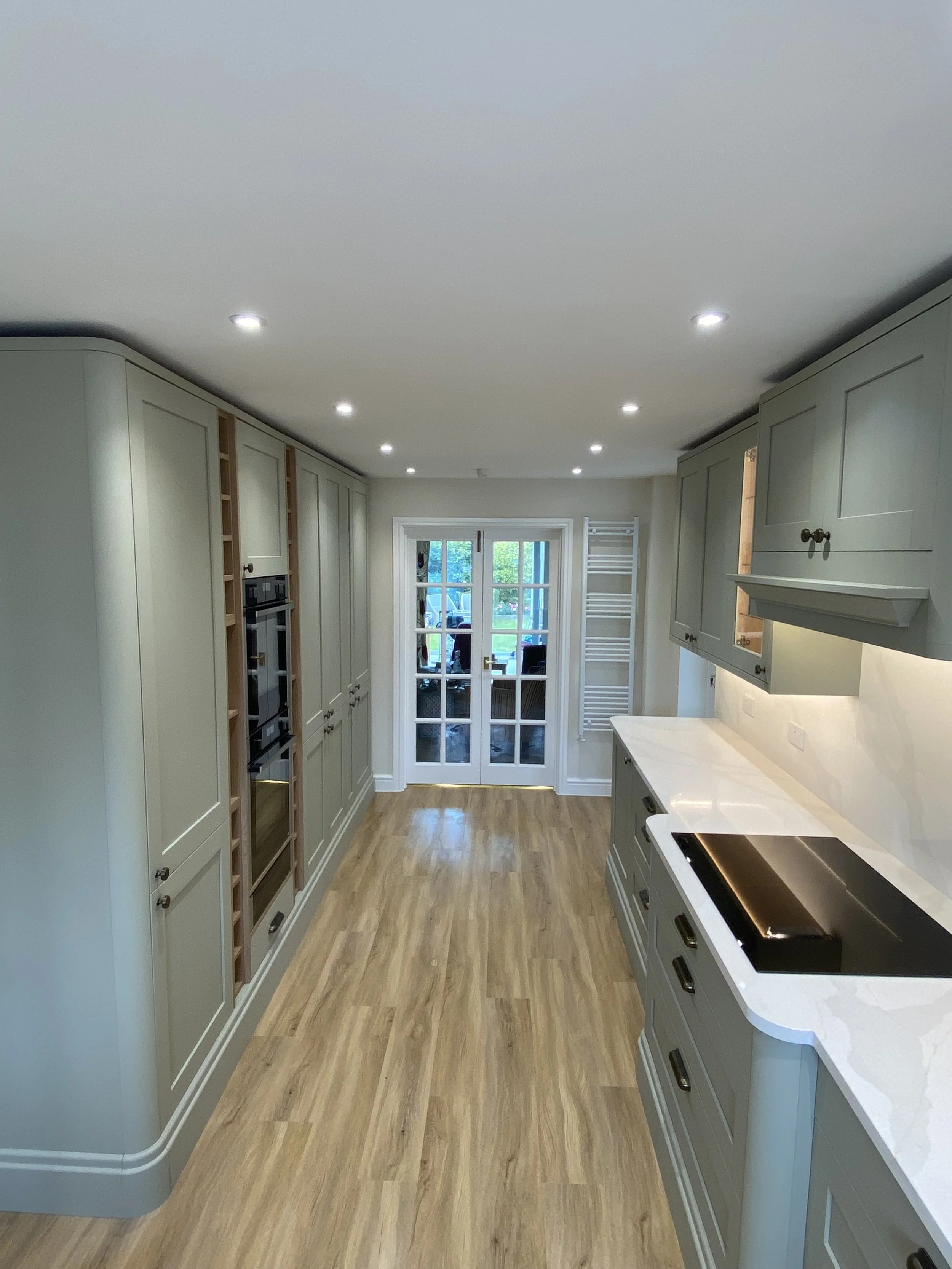 A modern kitchen with green cabinets, a white marble countertop, built-in appliances, wooden flooring, and double glass doors leading outside.