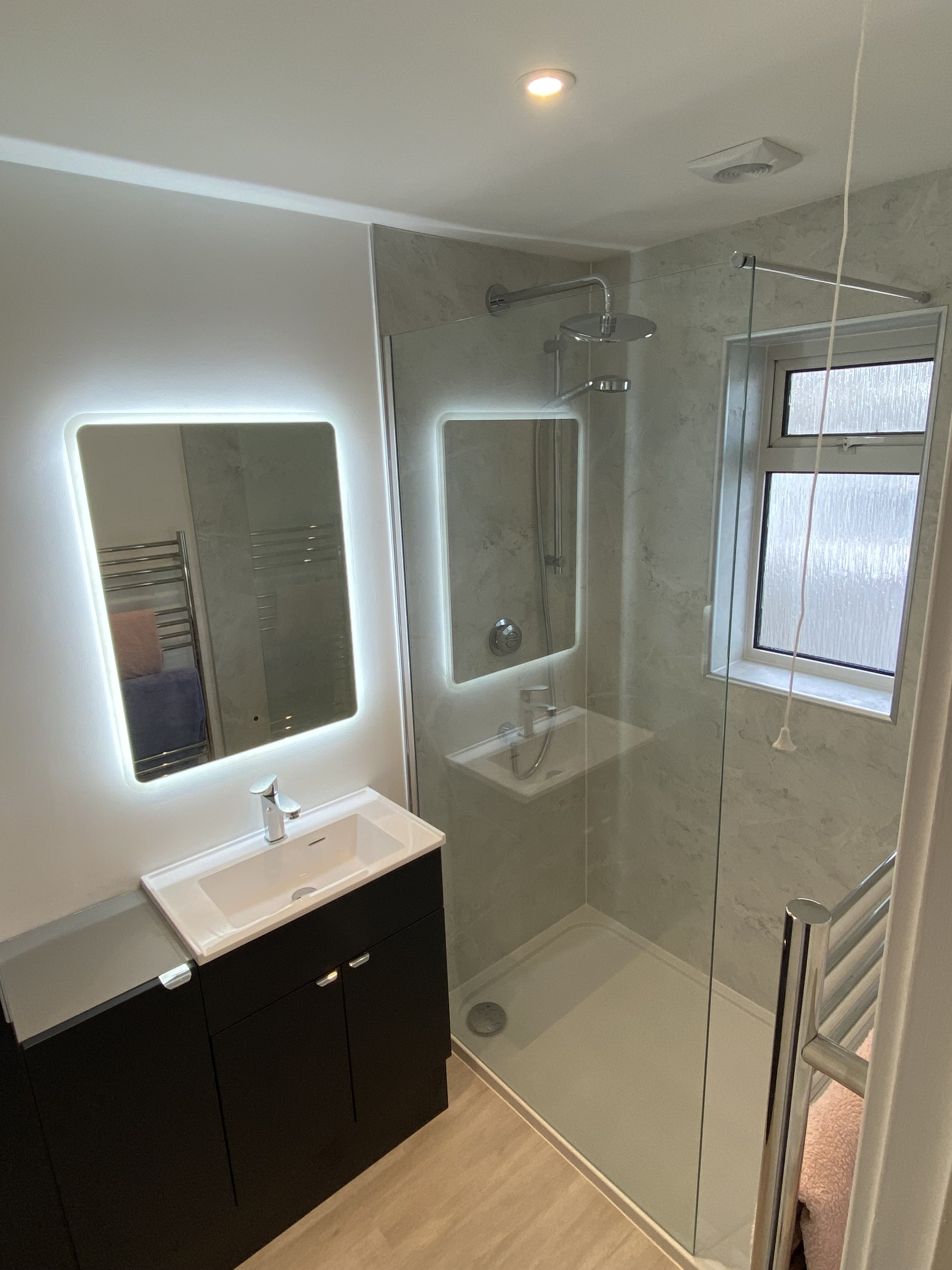 Black bathroom cupboard with white sink above. Rectangle mirror on the wall with lights surrounding. Wide shower with glass screen and window in the wall