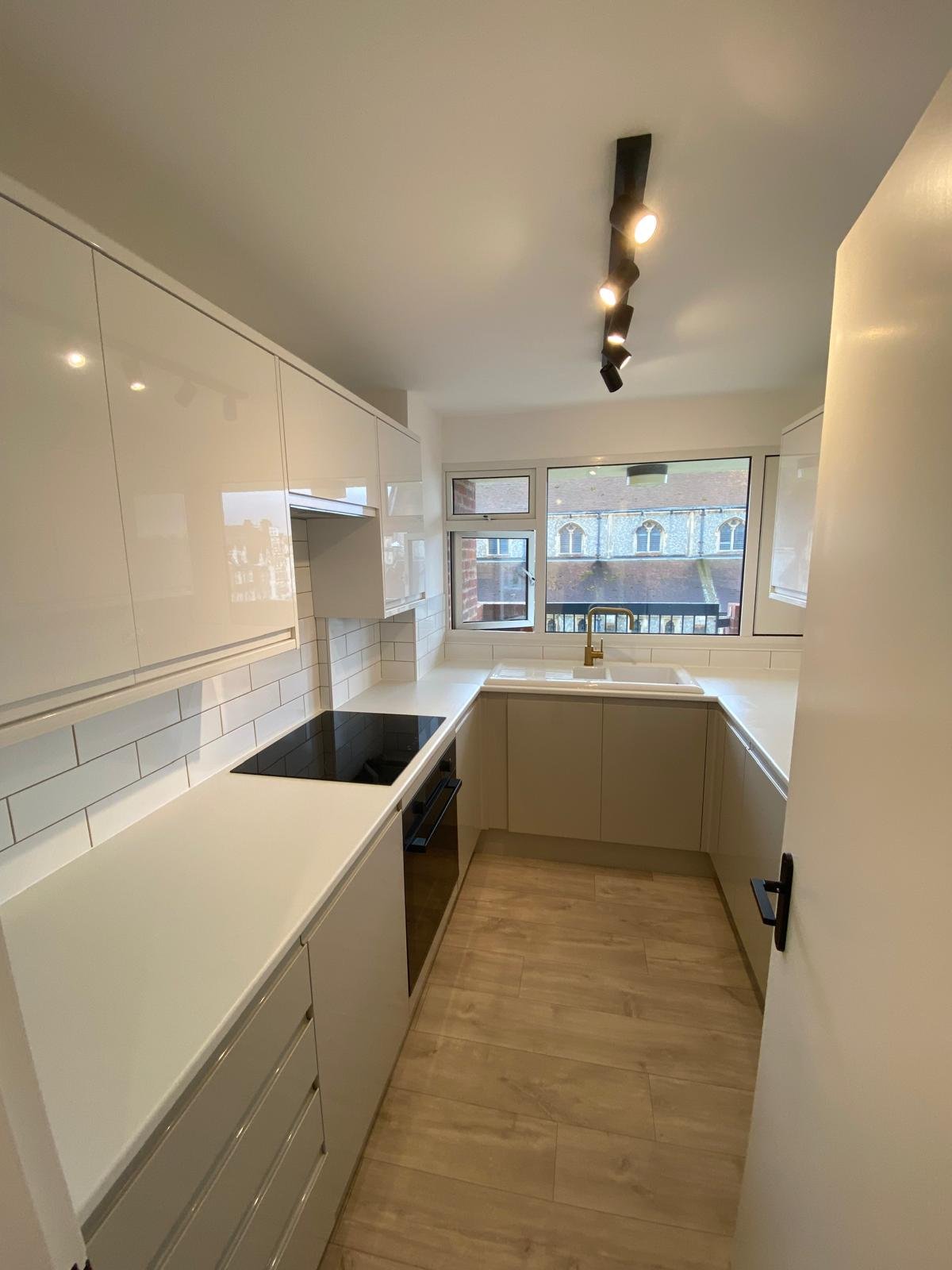 Small, modern kitchen with white cabinets, black stovetop, white countertop, and wooden flooring. There is a window above the sink with a view of brick buildings outside.