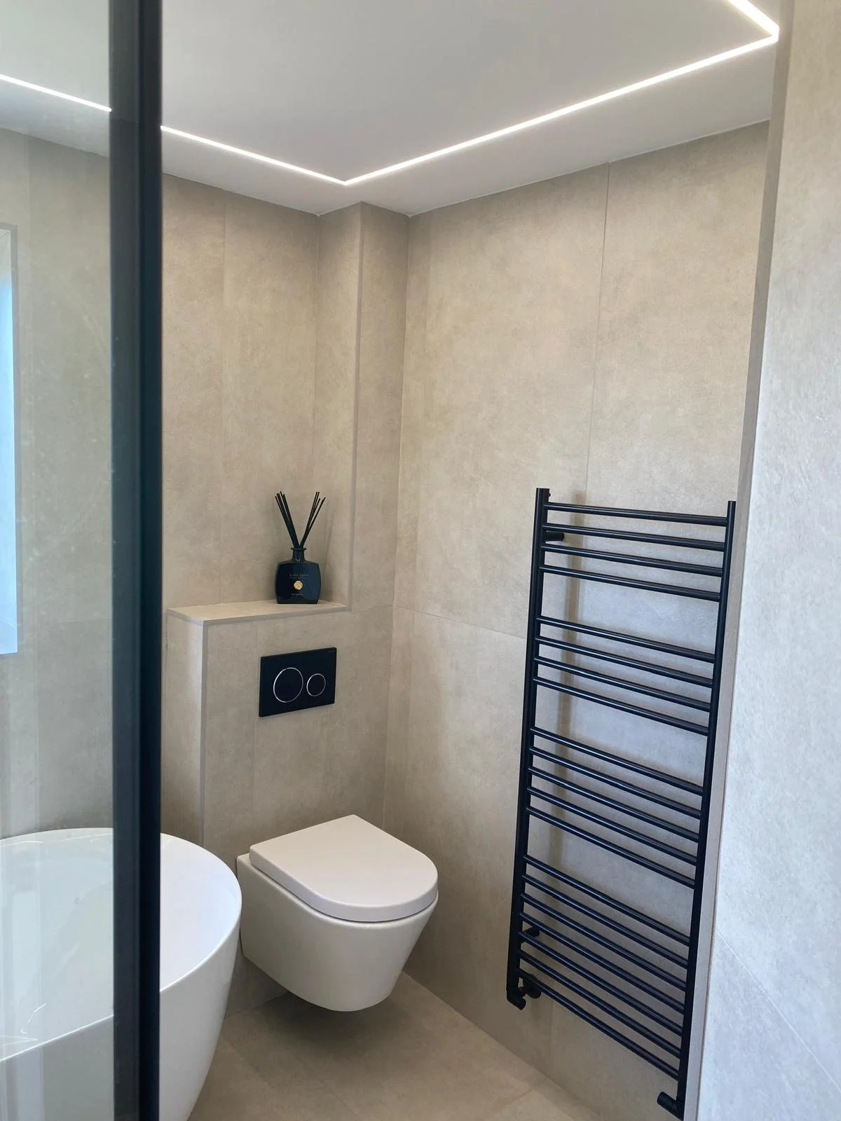 Beige tiled bathroom with toilet installed into wall with black flush buttons. Black full length radiator on wall