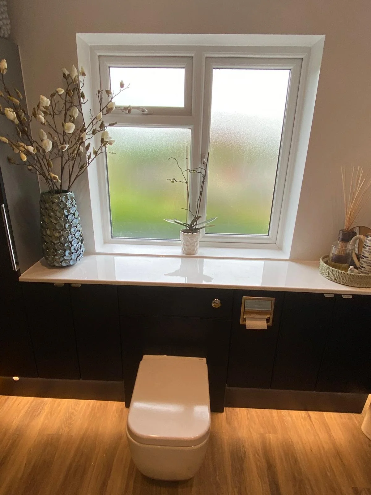A bathroom with a toilet in front of a window. The window has frosted glass, and there are decorative plants on the windowsill and in a large blue vase on the left side. To the right, there's a tray with toiletries. The countertop is white, and there