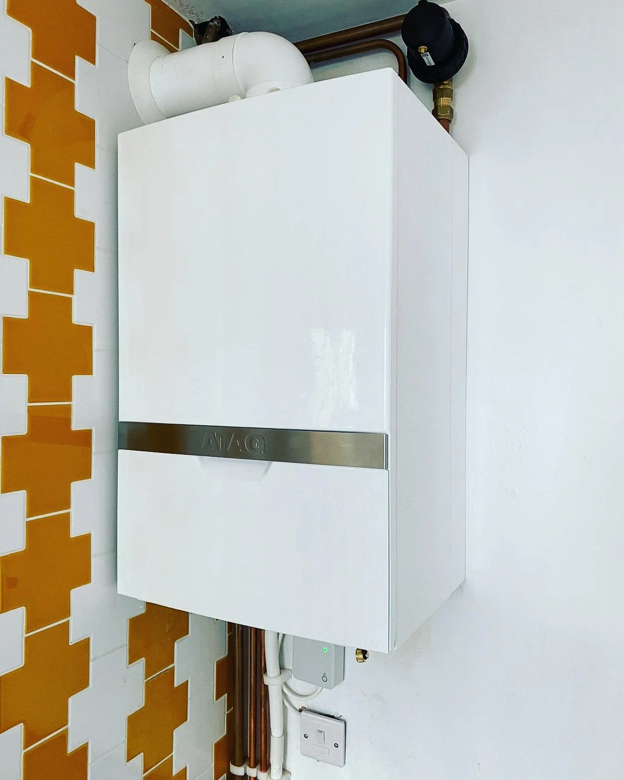 A white boiler system mounted on a wall with piping and a black component on top, located next to orange and white patterned tiles.