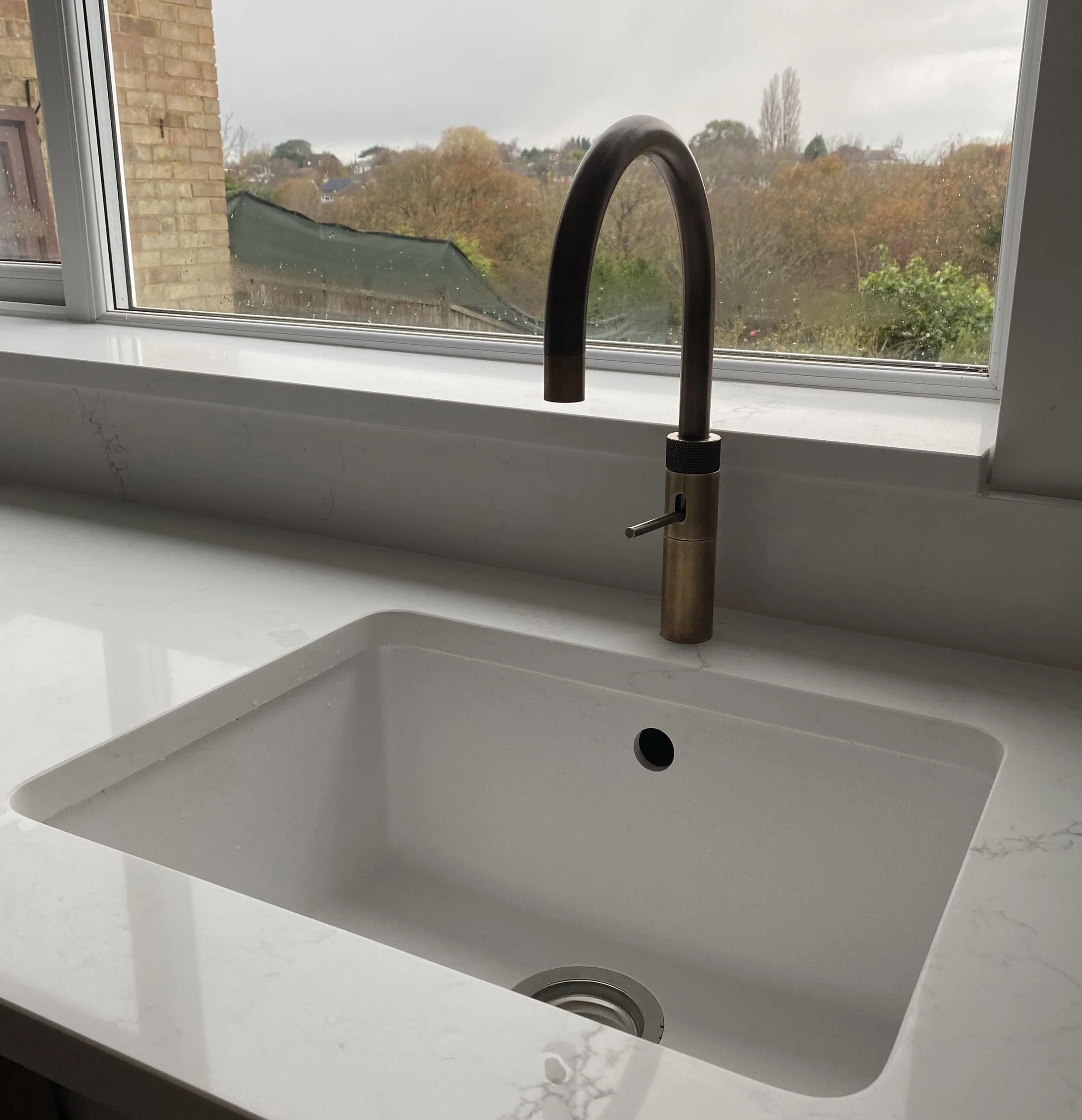 A kitchen sink with a modern brass faucet positioned in front of a window overlooking a neighborhood with trees and houses.