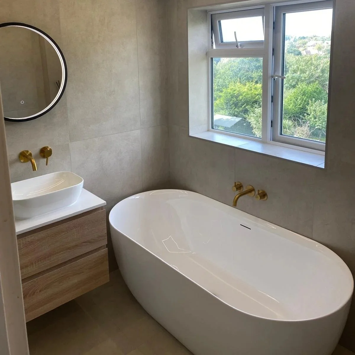 Modern bathroom with a freestanding bathtub, a small vanity with a vessel sink, a round mirror, and a window showing a view of trees.