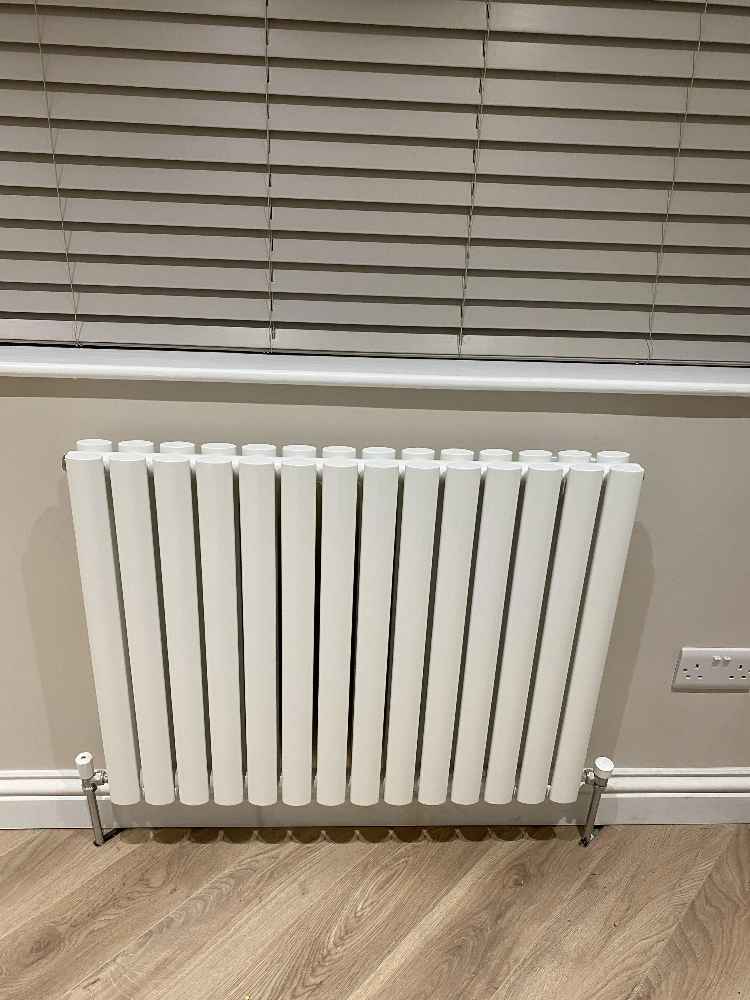 White radiator heater below a window with closed blinds, electrical outlet on the wall to the right, wooden floor.