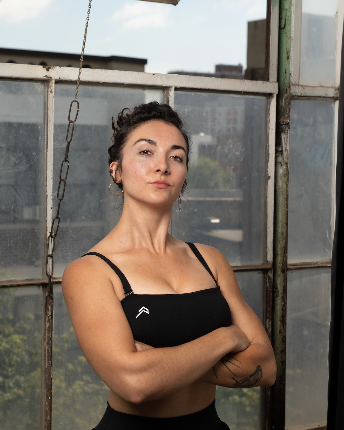 Confident woman with crossed arms and earrings posing in front of a weathered glass wall with city buildings in the background.