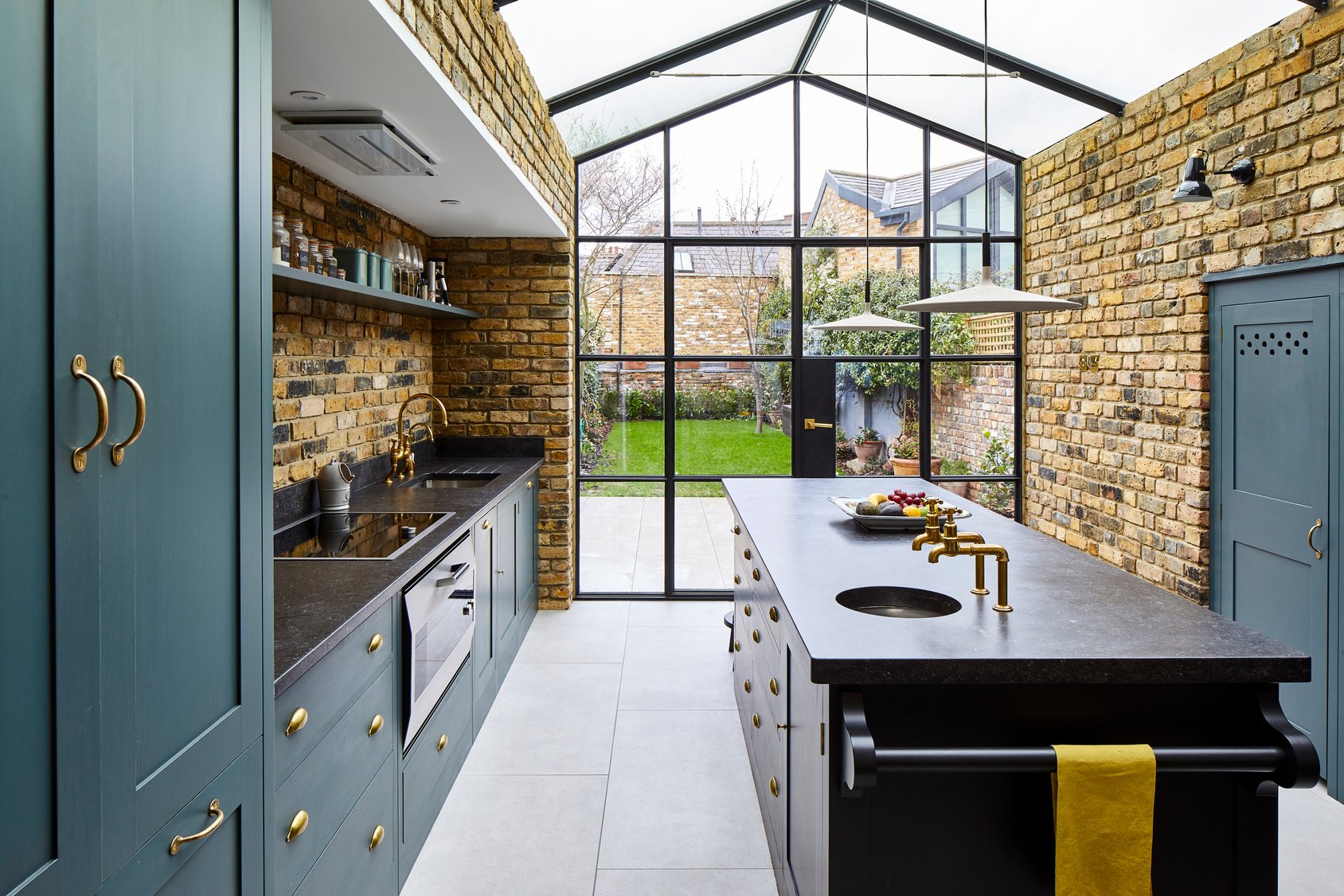 Stylish kitchen extension with exposed brick walls, vaulted glass ceiling, and Crittall-style doors leading to a garden. The space features brass fixtures, deep green cabinetry, and a central island with black countertops, blending industrial and cla