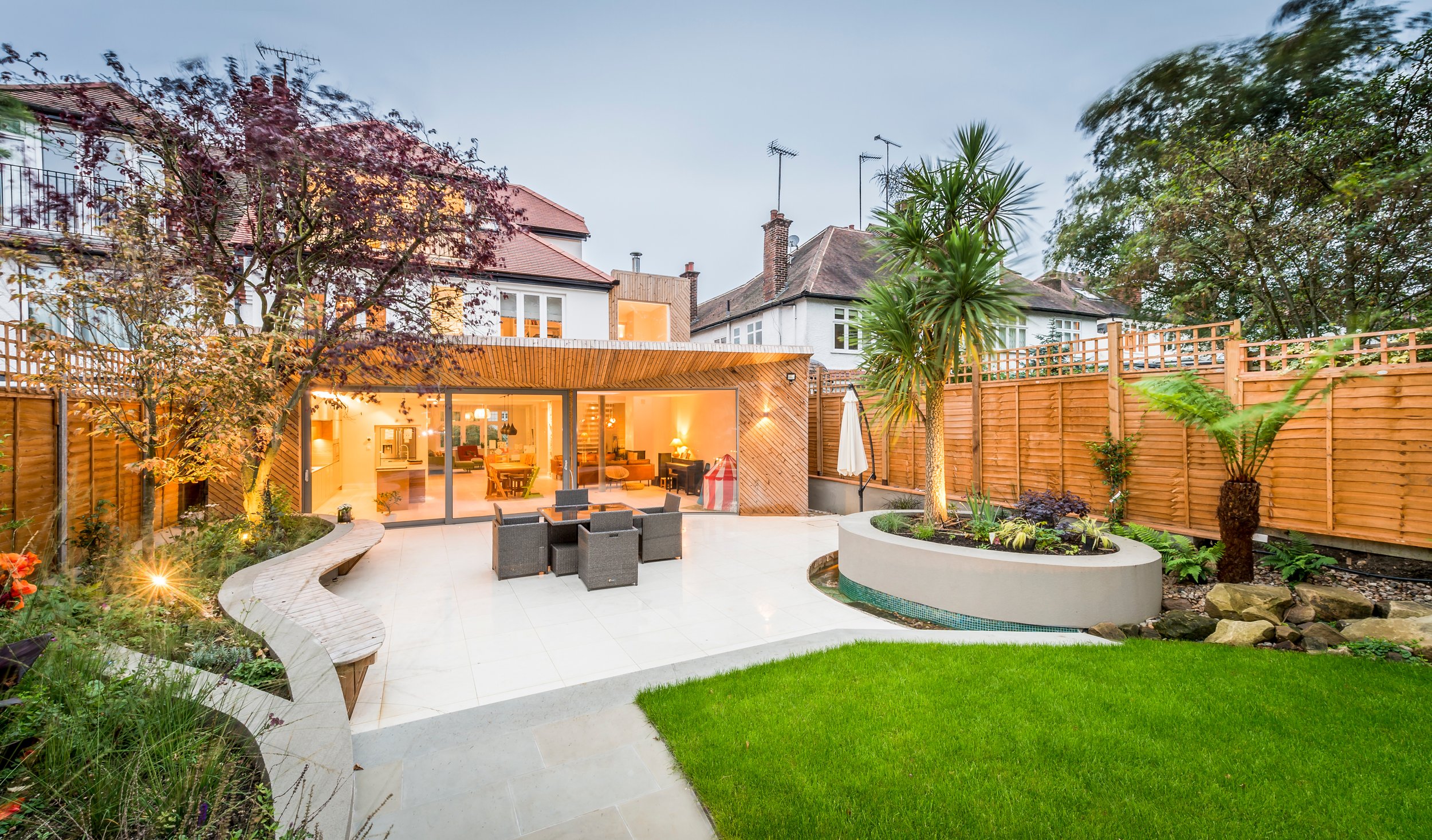 Backyard with outdoor seating, garden beds, trees, and a house with large sliding glass doors, illuminated in the evening.