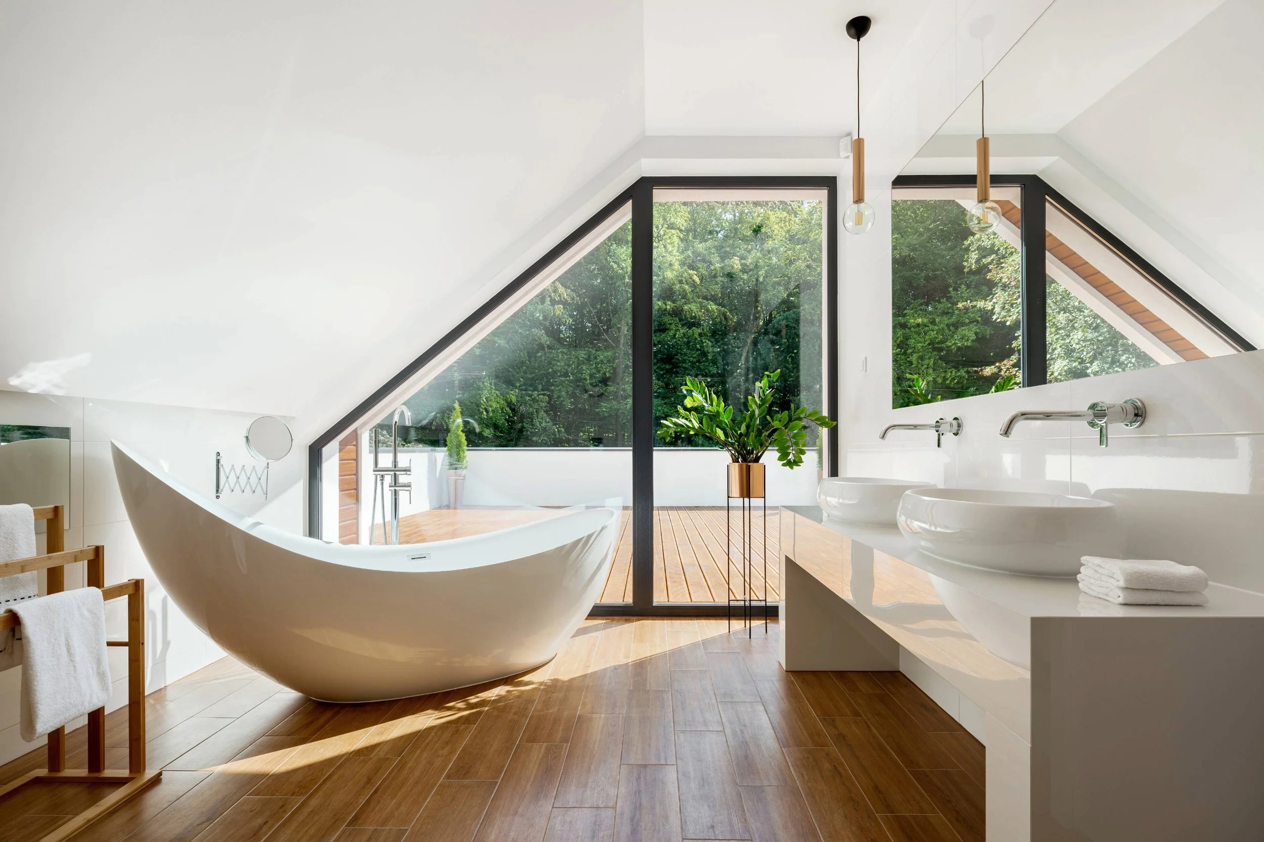 Luxury attic bathroom featuring a sculptural freestanding bath, dual basins, and floor-to-ceiling glazing with views of a private terrace and surrounding trees, blending modern design with natural light and serene ambience