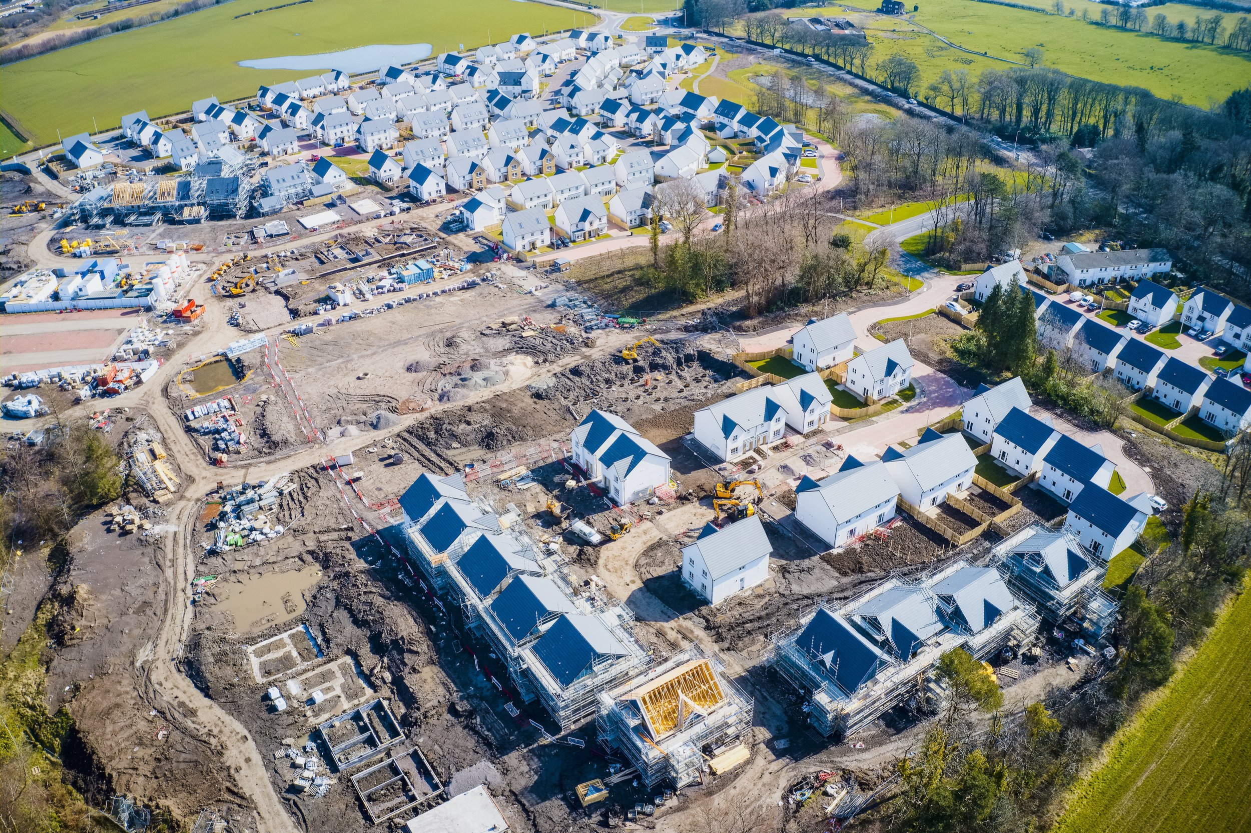 Aerial view of a large-scale new housing development under construction, showing partially built homes, foundations, and construction equipment alongside completed residential units, representing modern UK housing expansion