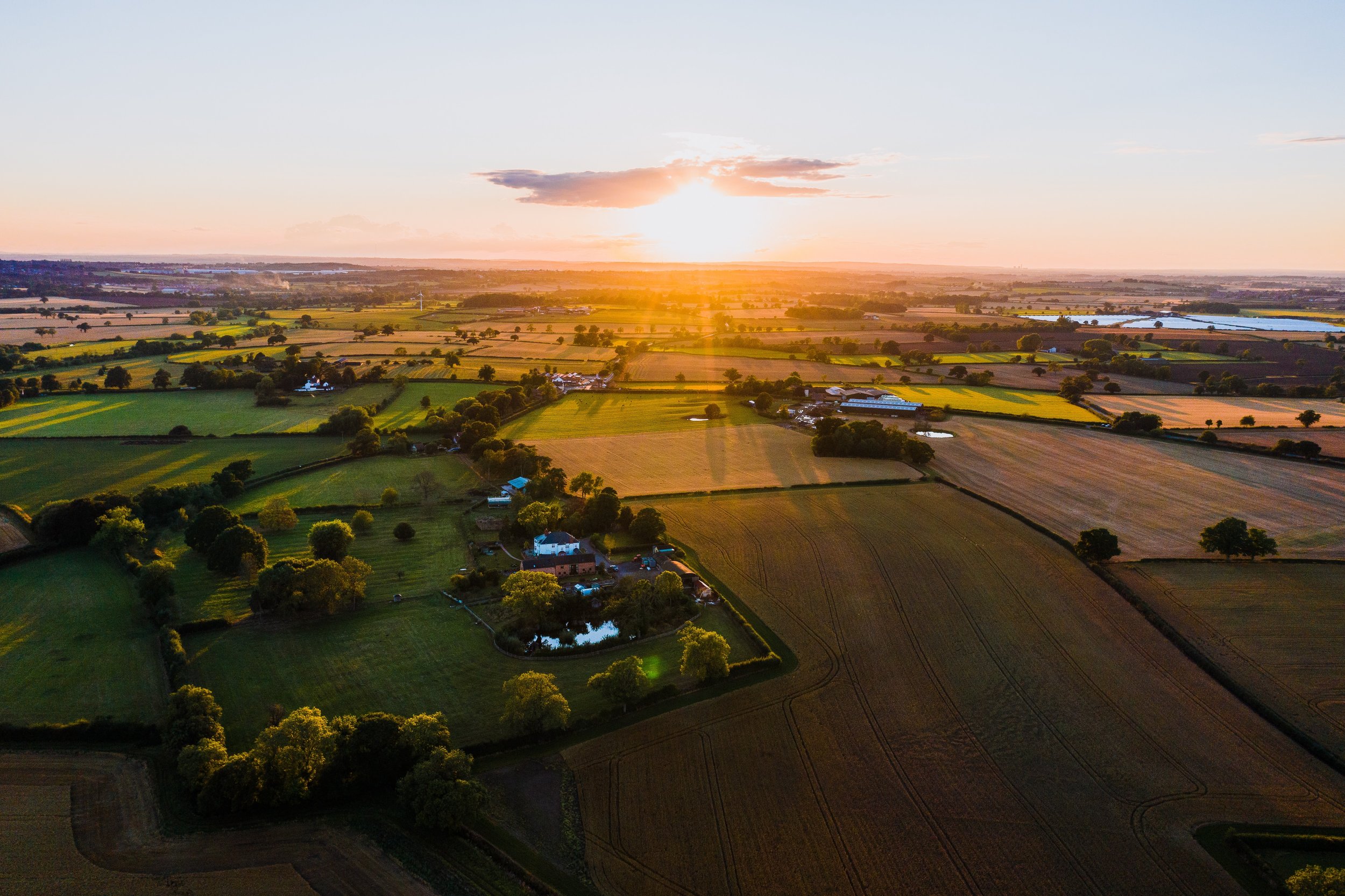 Aerial view of UK countryside at sunset, with golden light casting long shadows across patchwork fields, hedgerows, and rural farmland, capturing the beauty and tranquillity of the British landscape