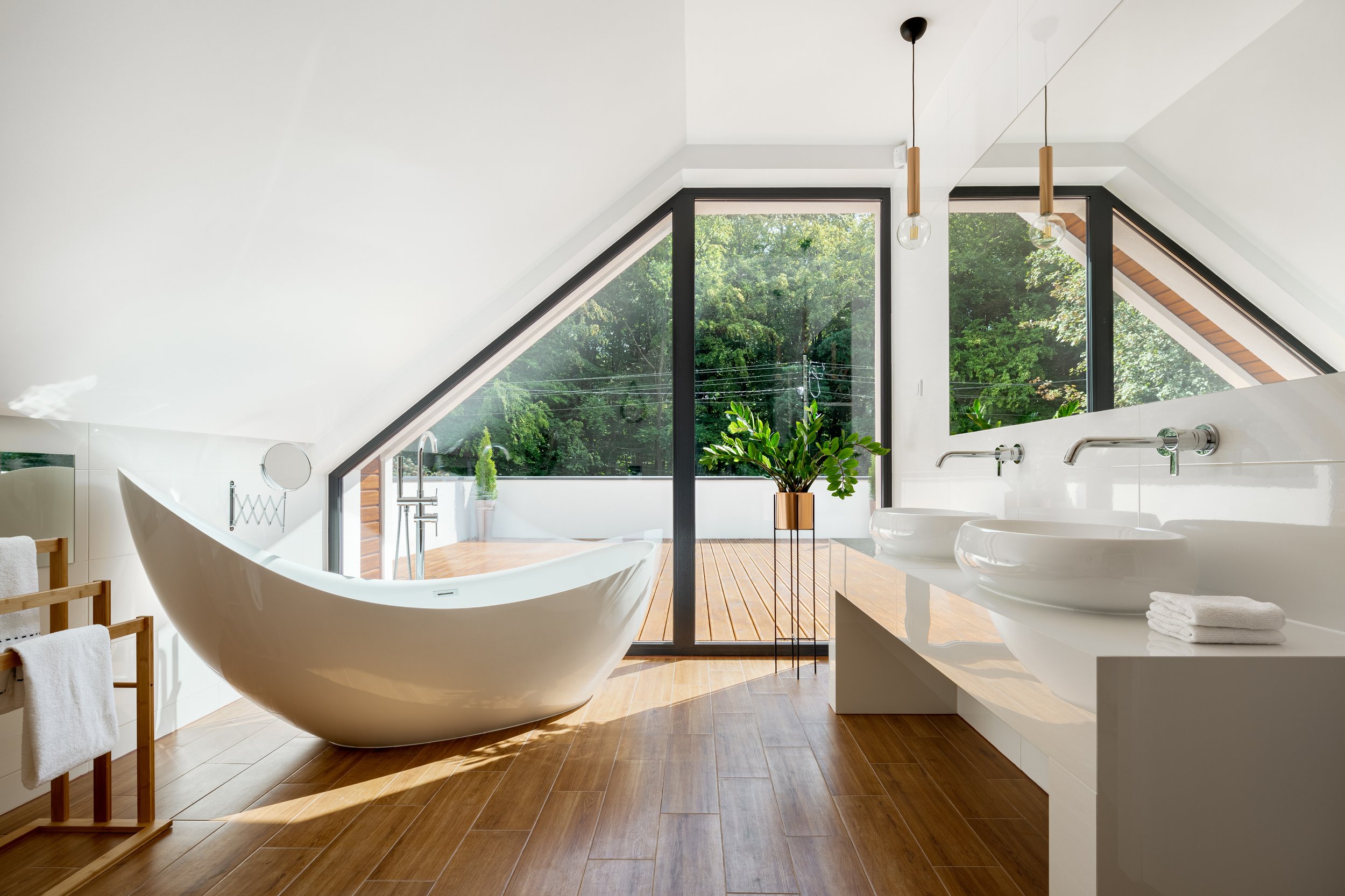 Luxury attic bathroom featuring a sculptural freestanding bathtub, dual basins, and floor-to-ceiling glazing with views of a private terrace and surrounding trees, blending modern design with natural light and serene ambience