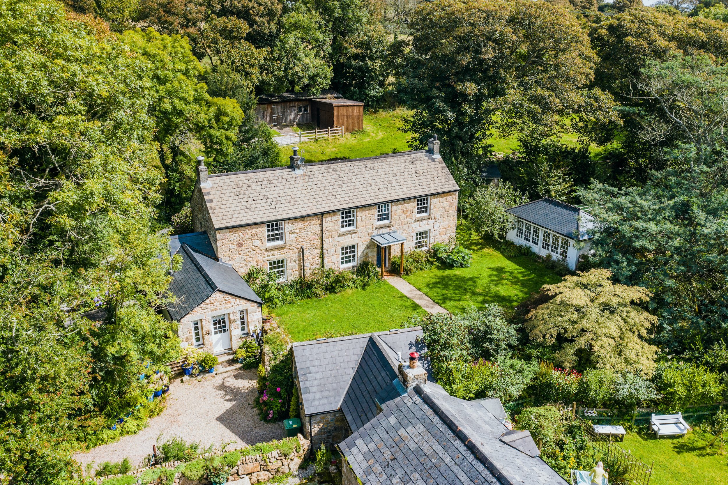 Aerial view of a traditional stone cottage surrounded by mature trees, landscaped gardens, and outbuildings — a charming rural UK property captured in bright daylight from above