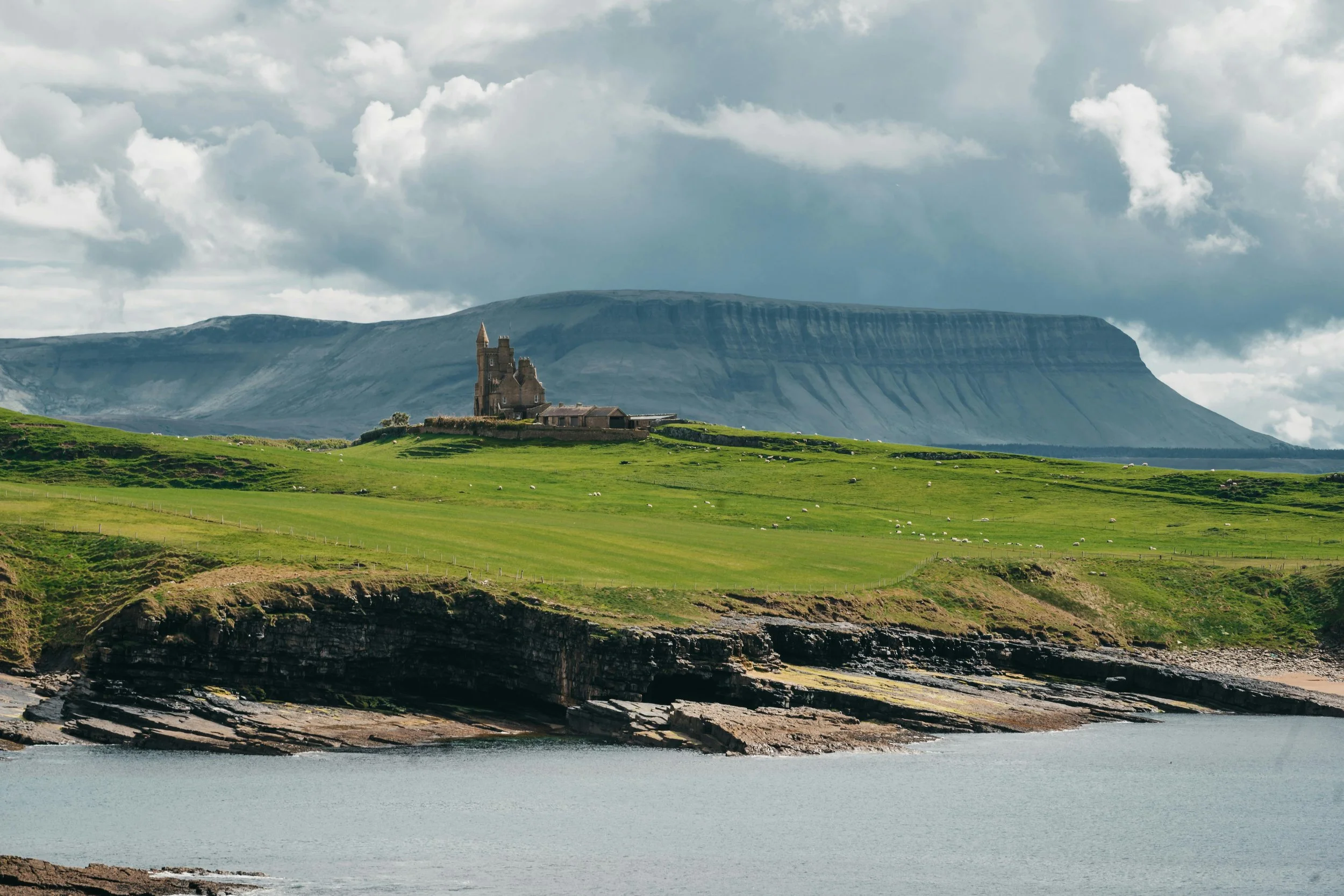 Castle on a grassy hill with a mountain background, overlooking a rocky coastline and sea.