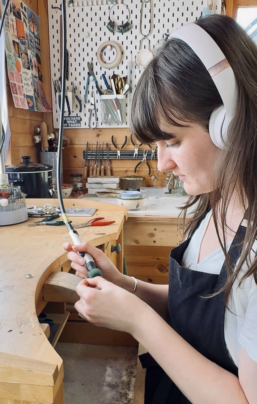 A woman working on a woodworking project in a workshop. She is wearing a headband and headphones, focusing on shaping a piece of wood with a tool. The workspace is equipped with various tools, scissors, and organized supplies on pegboards and shelves.