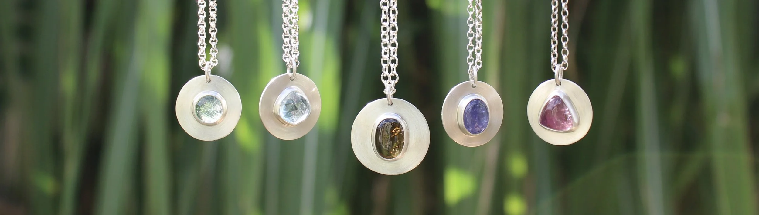 Five silver necklaces with oval-shaped colored gemstones hanging in front of green foliage.