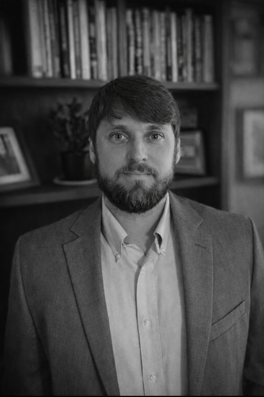 A black-and-white portrait of a man with a beard and mustache, wearing a blazer over a button-up shirt, standing in front of a bookshelf with framed photos and a plant.