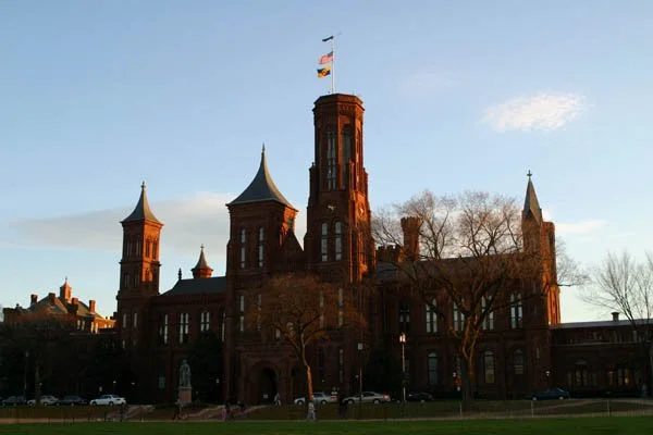 The red sandstone Smithsonian Castle and its towers seen across a lawn beneath a blue sky. Archival photograph by Thunk, November 2002.