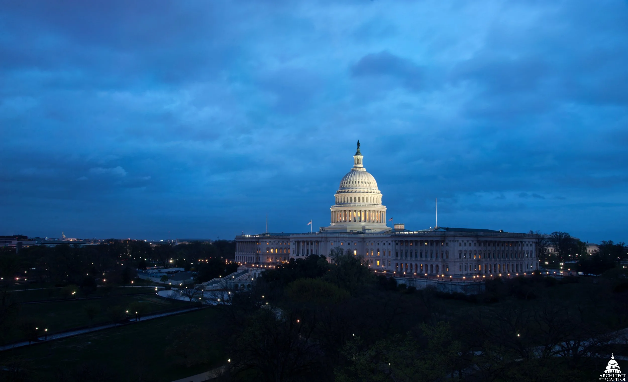 Archival view of the illuminated U.S. Capitol dome and west front beneath a blue evening sky, with dark trees and grounds in the foreground. Architect of the Capitol photograph, April 2014.