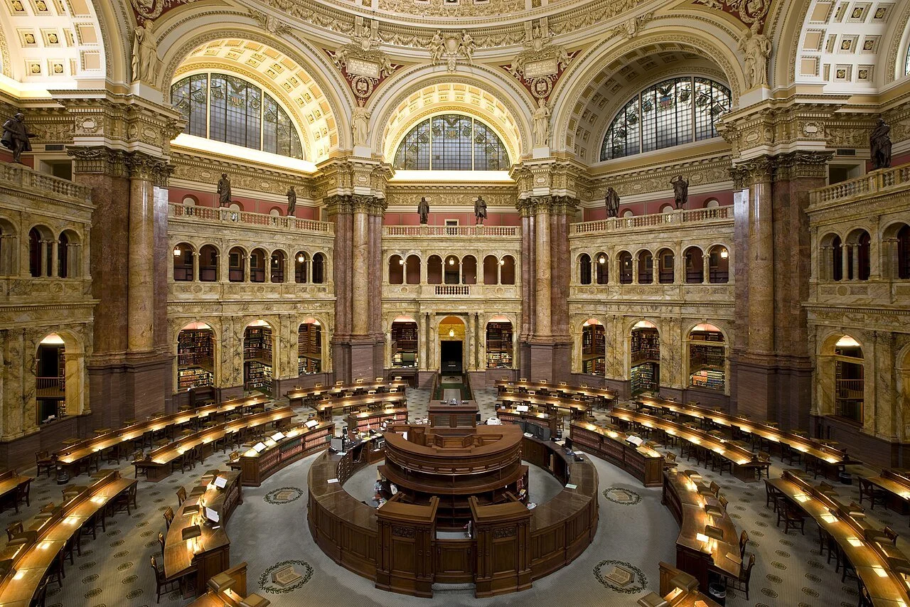 The Library of Congress Main Reading Room, with curved rows of wooden desks beneath tall marble columns and arched windows. Archival photograph by Carol M. Highsmith, 2009.