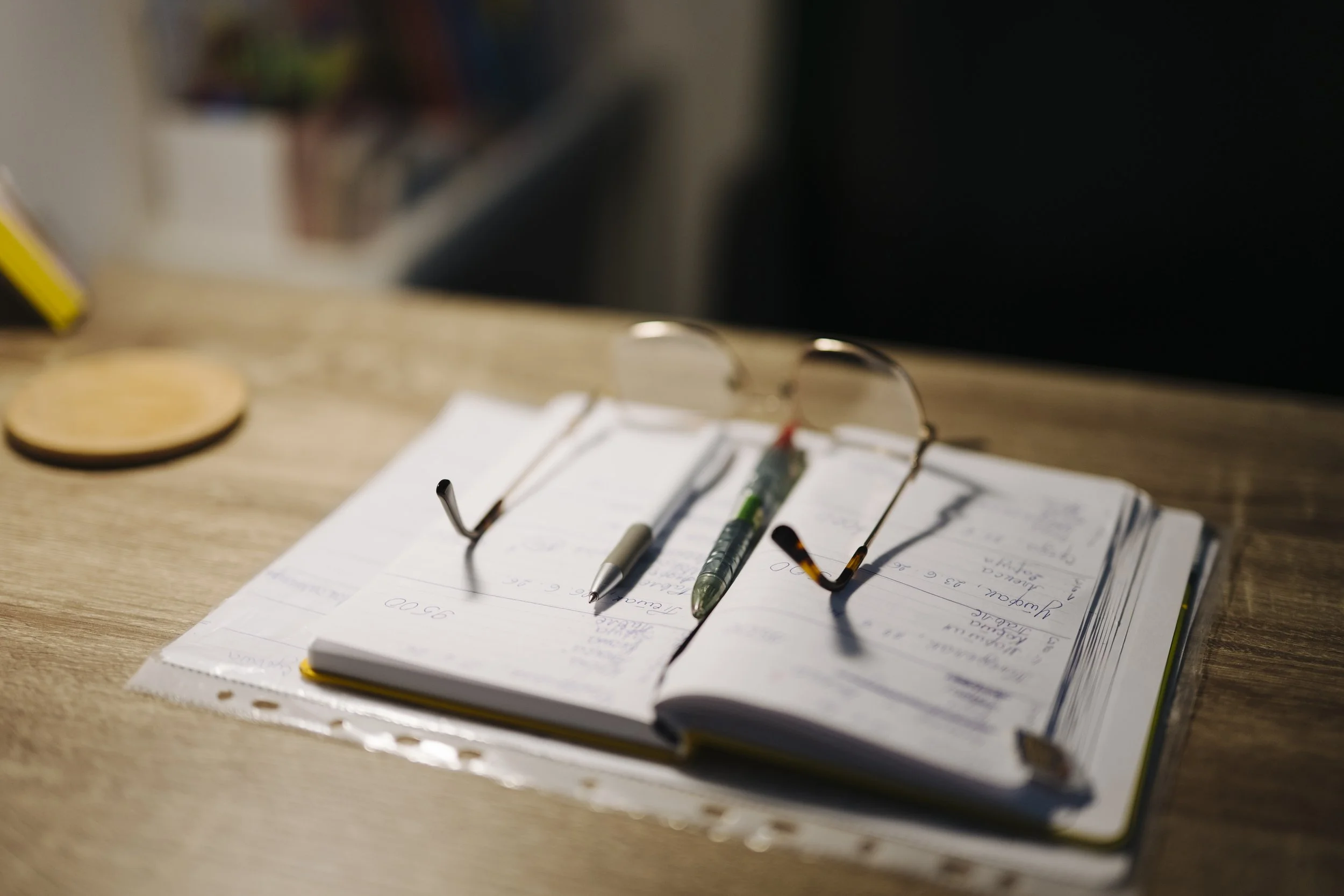 Reading glasses and two pens rest across an open notebook on a wooden desk. Illustrative photograph, June 27, 2026.