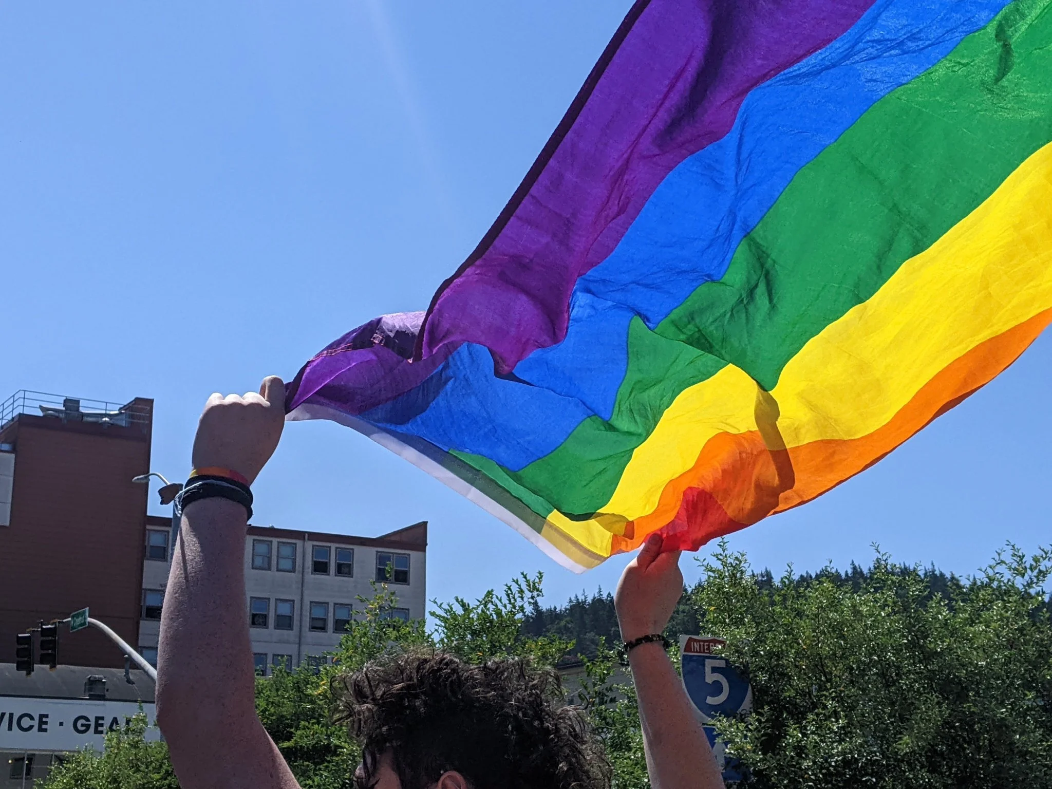 A person holds a large rainbow flag overhead against a blue sky at the Bellingham Pride Parade in July 2023. Archival photograph by Robert Ashworth.