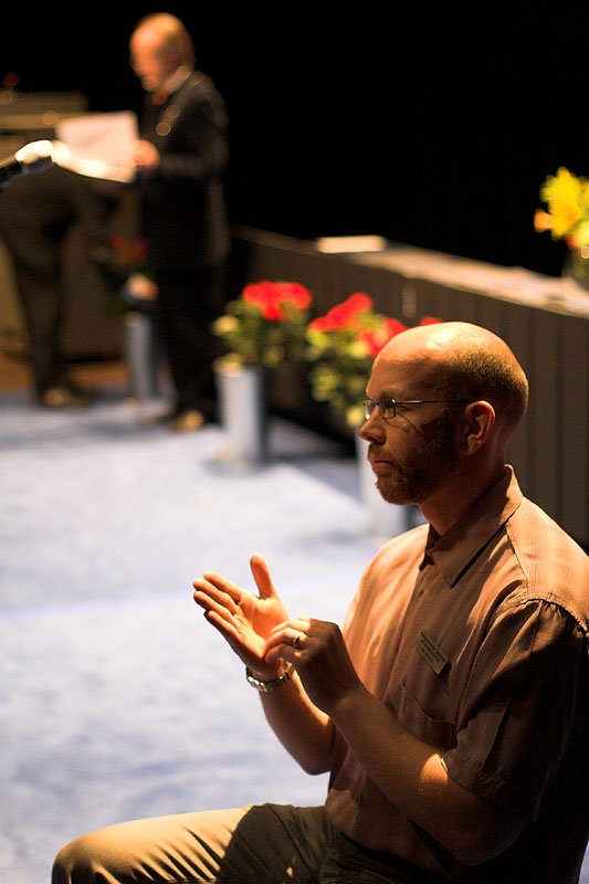 A seated interpreter signs with both hands in the foreground while a speaker stands at a lectern behind him. Archival photograph from May 2005.