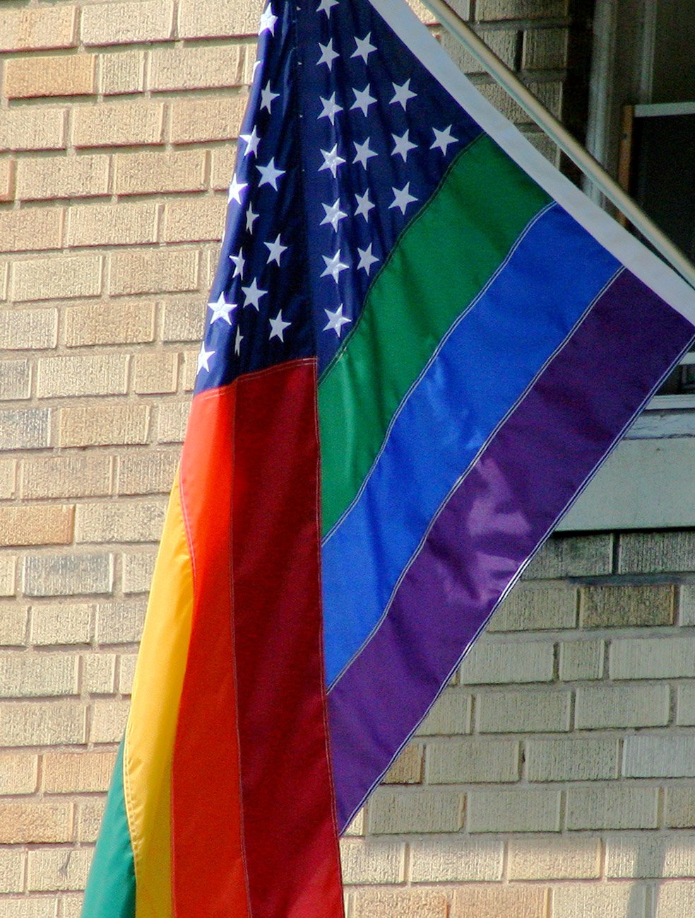 Archival illustrative photo of a rainbow-striped flag with white stars on a blue field hanging beside a brick wall. Photo by dbking, 2006.