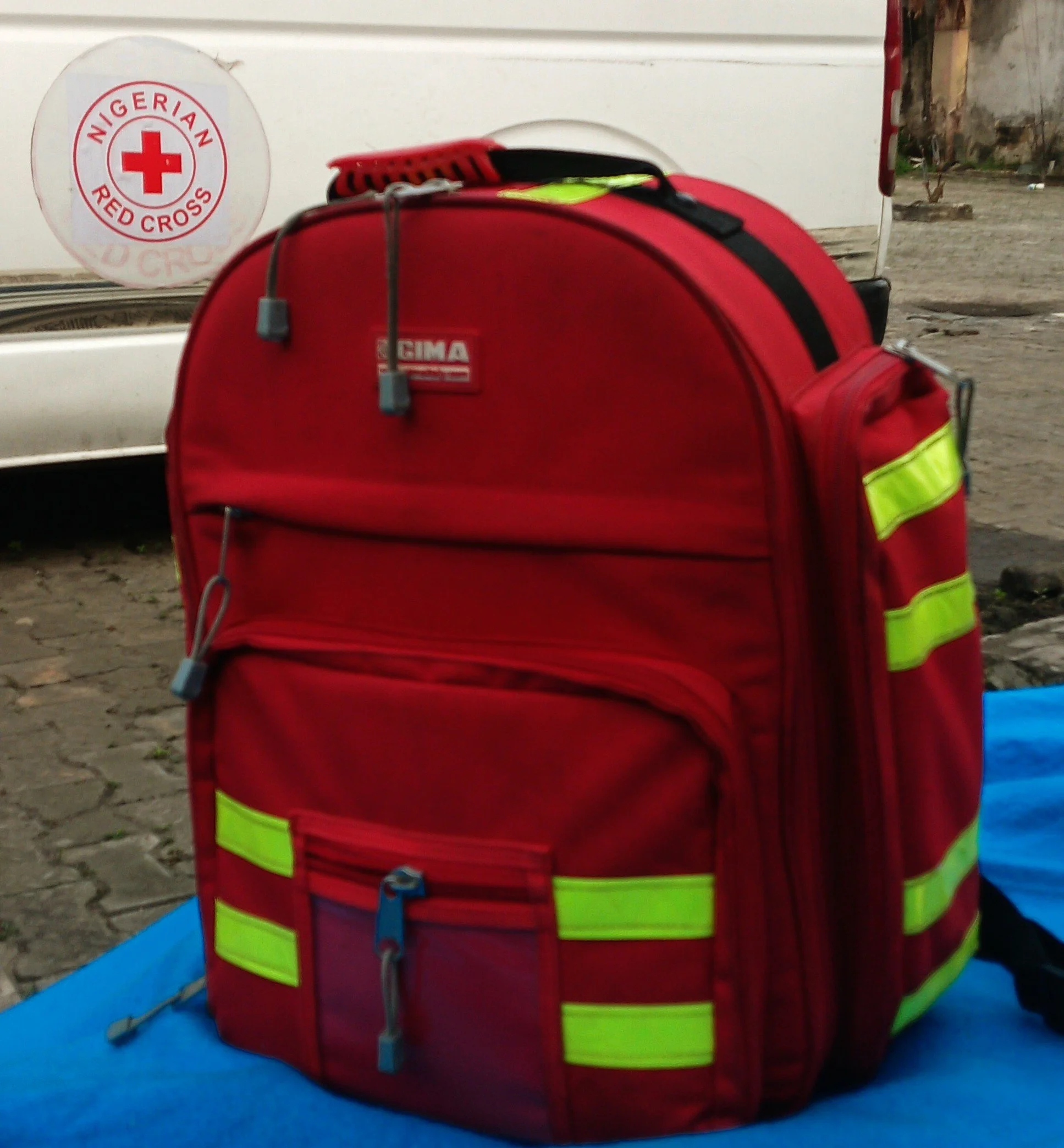Red first-aid backpack with bright reflective strips on a blue surface, with a Nigerian Red Cross emblem on a vehicle behind it. Archival illustrative photograph, December 2025.