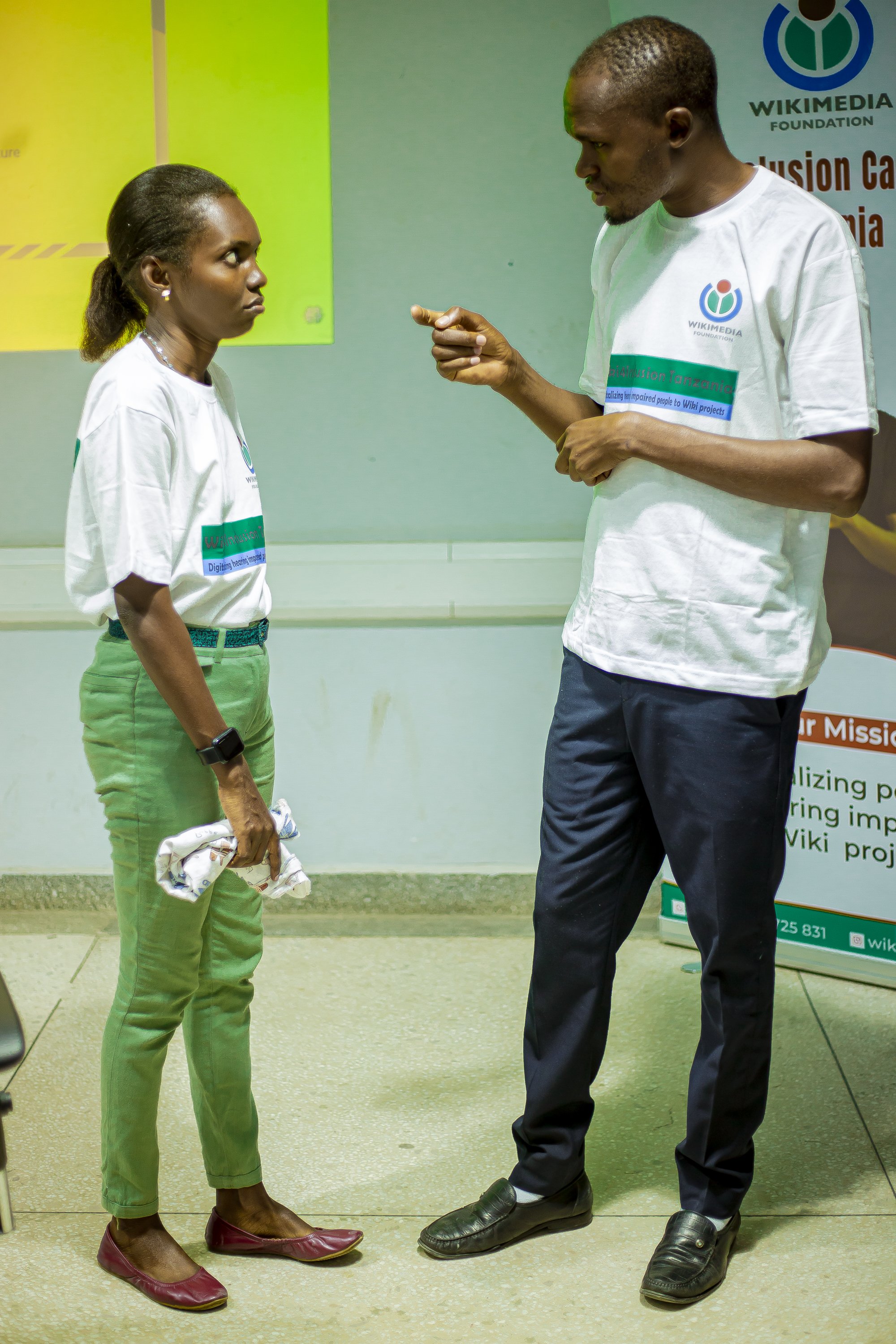 Two people facing one another at a Wiki4Inclusion training, one signing with raised hands. Archival photograph from Tanzania, July 2024; the source identifies Tanzanian Sign Language.