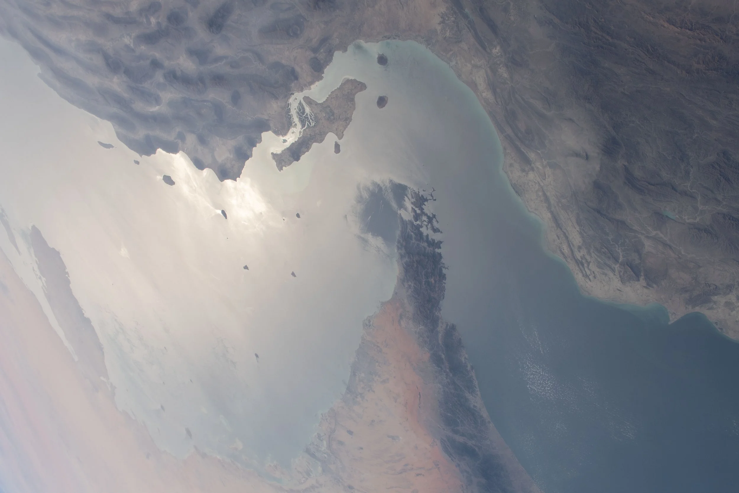 Archival orbital photograph of the Strait of Hormuz, showing pale rocky coastlines surrounding blue-gray water, with sunlight reflecting off the sea. NASA, March 24, 2021.