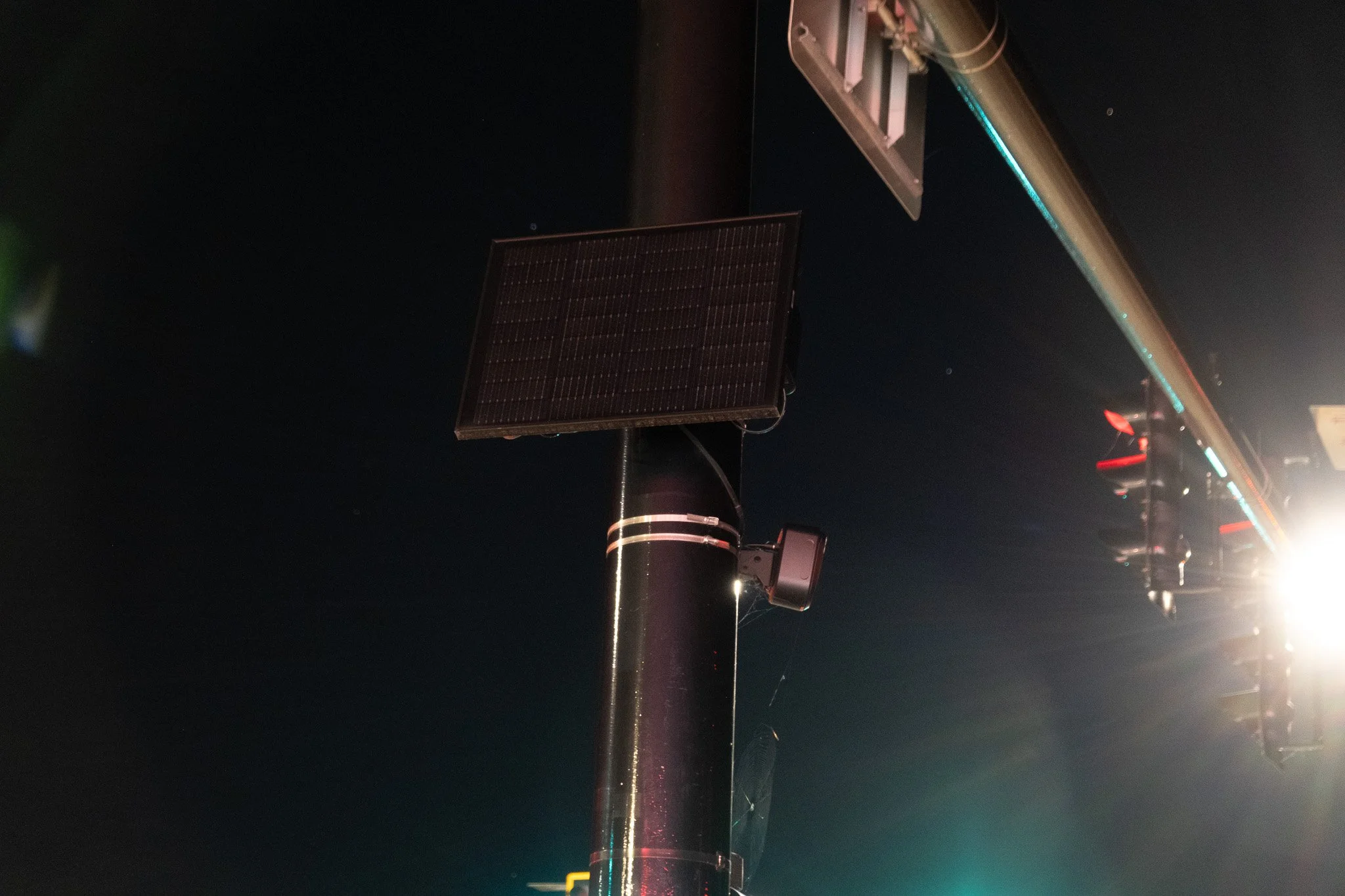 A Flock camera and rectangular solar panel mounted on a pole beside traffic lights at night in La Crosse, Wisconsin, August 23, 2026. Photo by Snoowastaken.