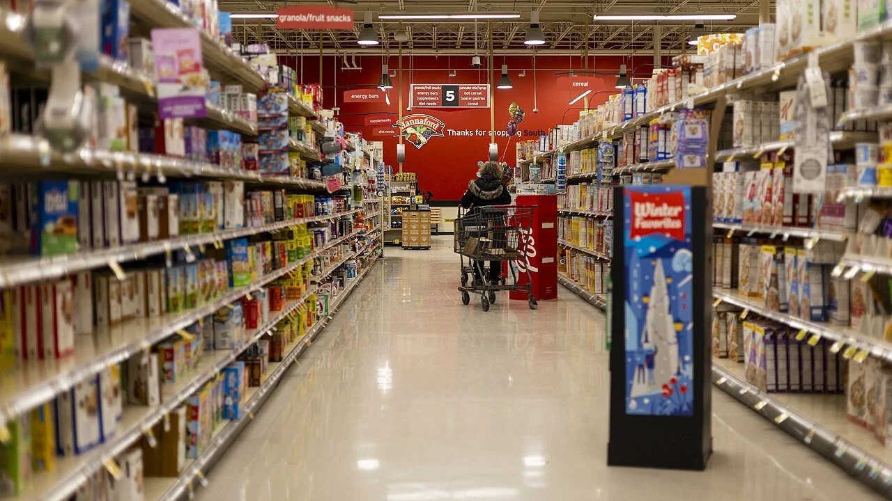 A stocked supermarket aisle with a shopping cart and a shopper near the far end, photographed in Vermont in December 2025.