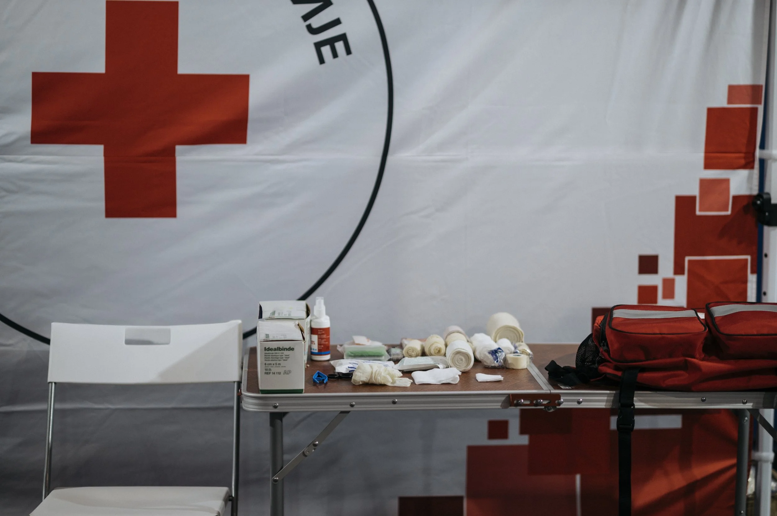 Bandages and first-aid supplies on a table beside red bags, with Red Cross banners behind them. Illustrative photograph from April 26, 2026.
