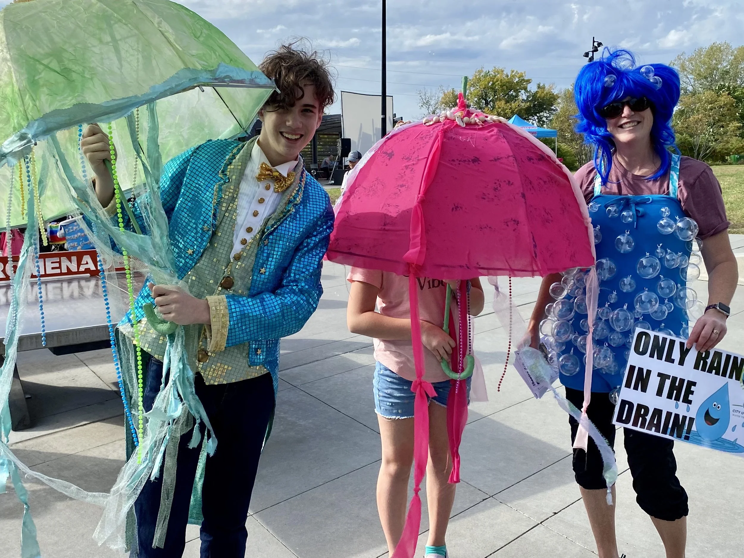 Rivers volunteers dressed in bright happy colors and outfits with umbrellas. One holds a sign "Only rain in the drain."