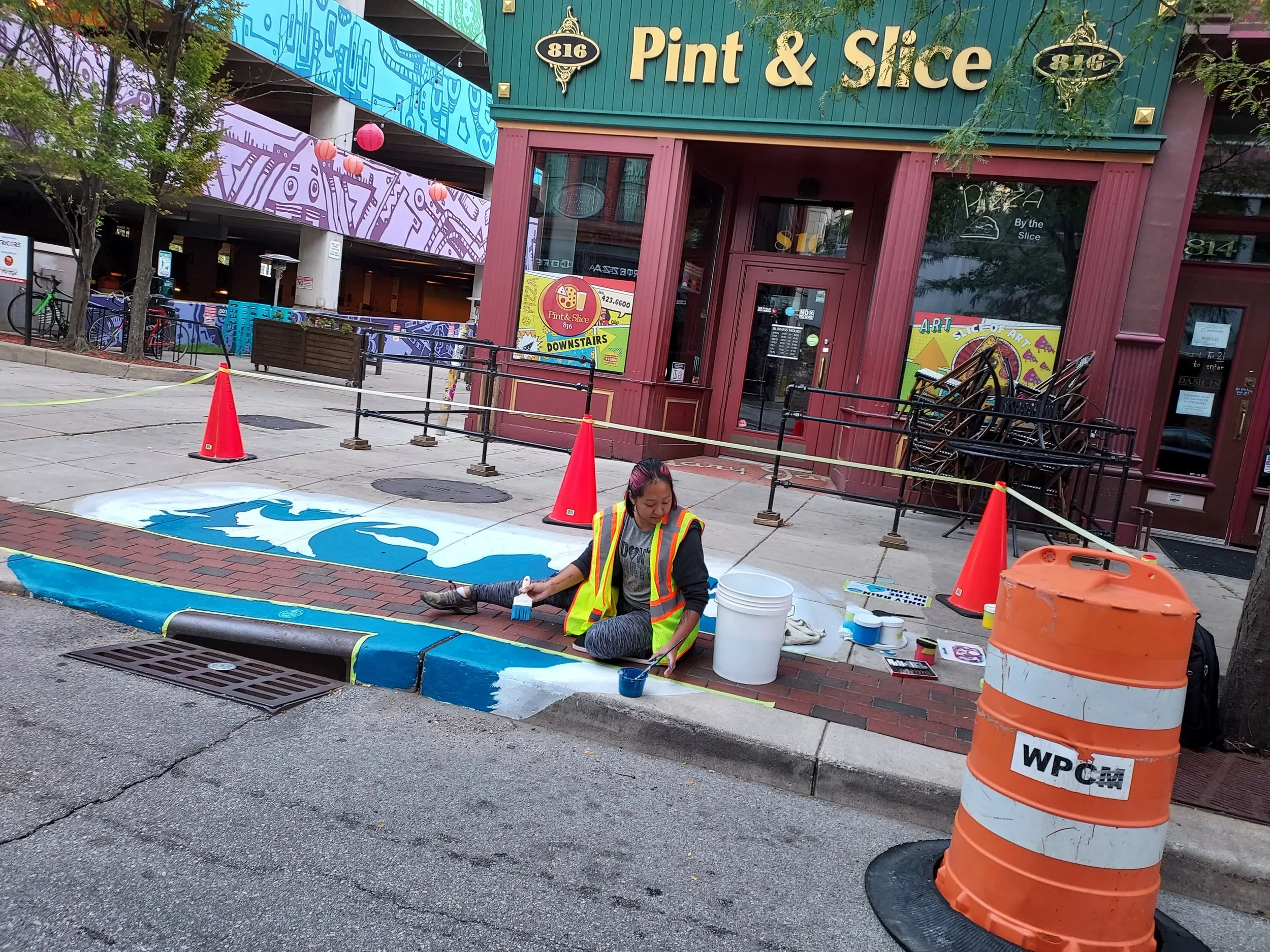 A women is painting the sidewalk by a drain outside Pint & Slice downtown Fort Wayne.