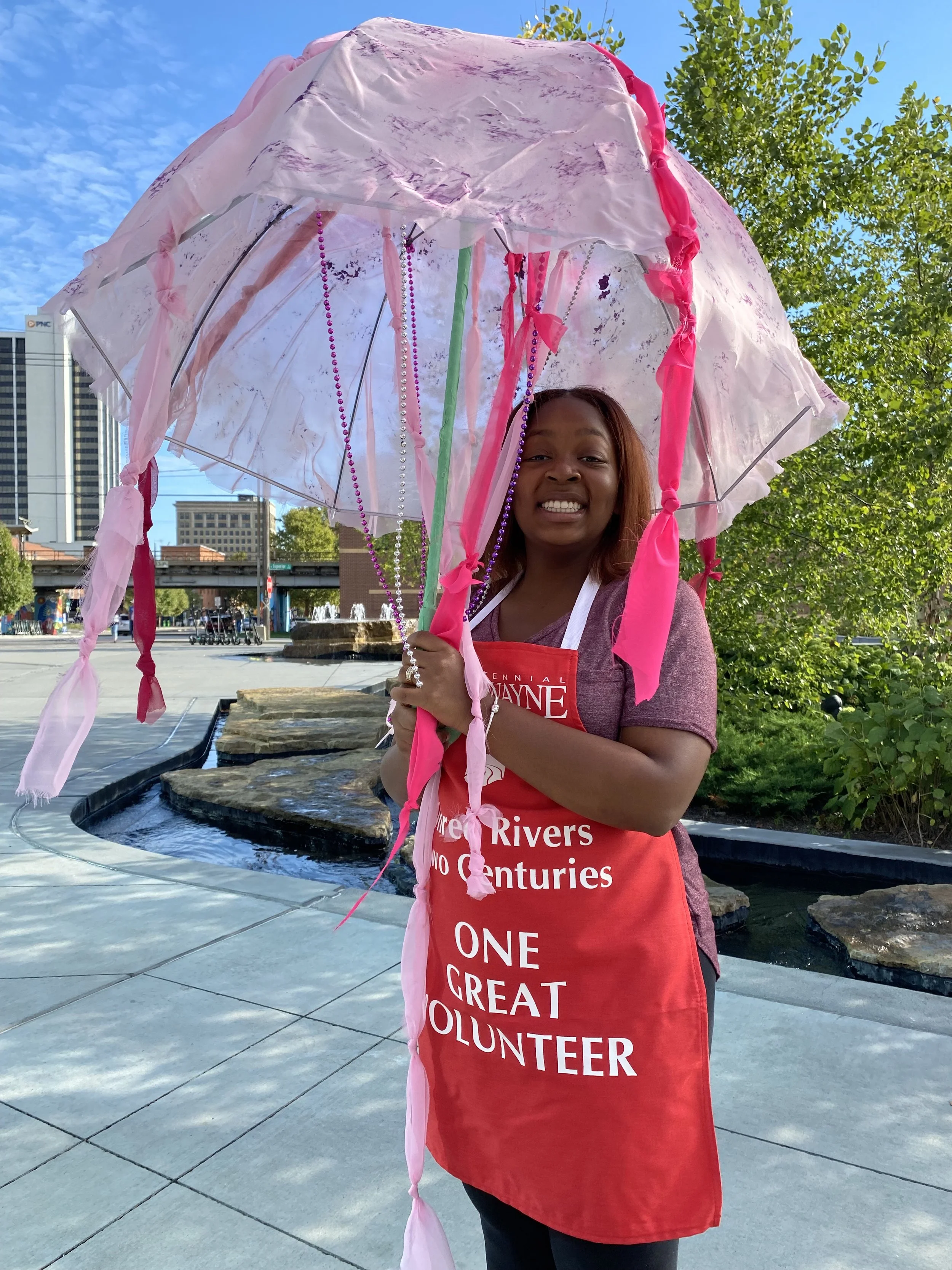 A smiling woman who is volunteering for the rivers, wearing a red apron and carrying an umbrella.