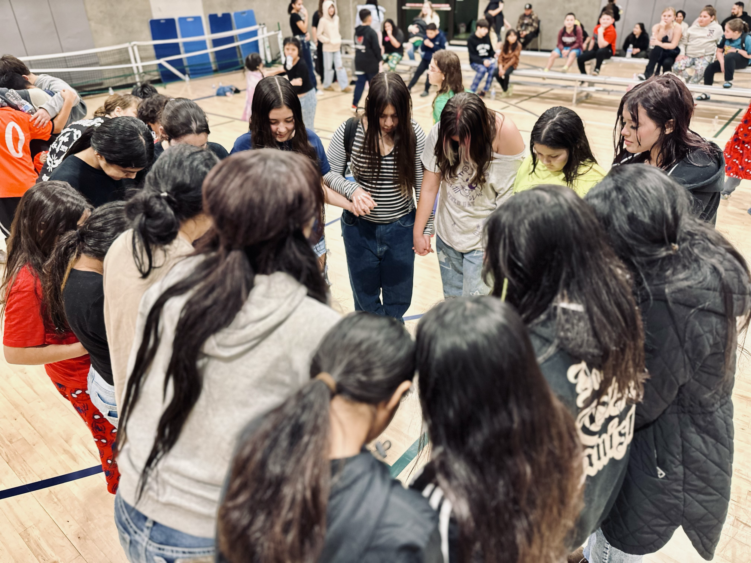 Group of people, mostly young women and girls, standing in a circle holding hands in an indoor gymnasium.