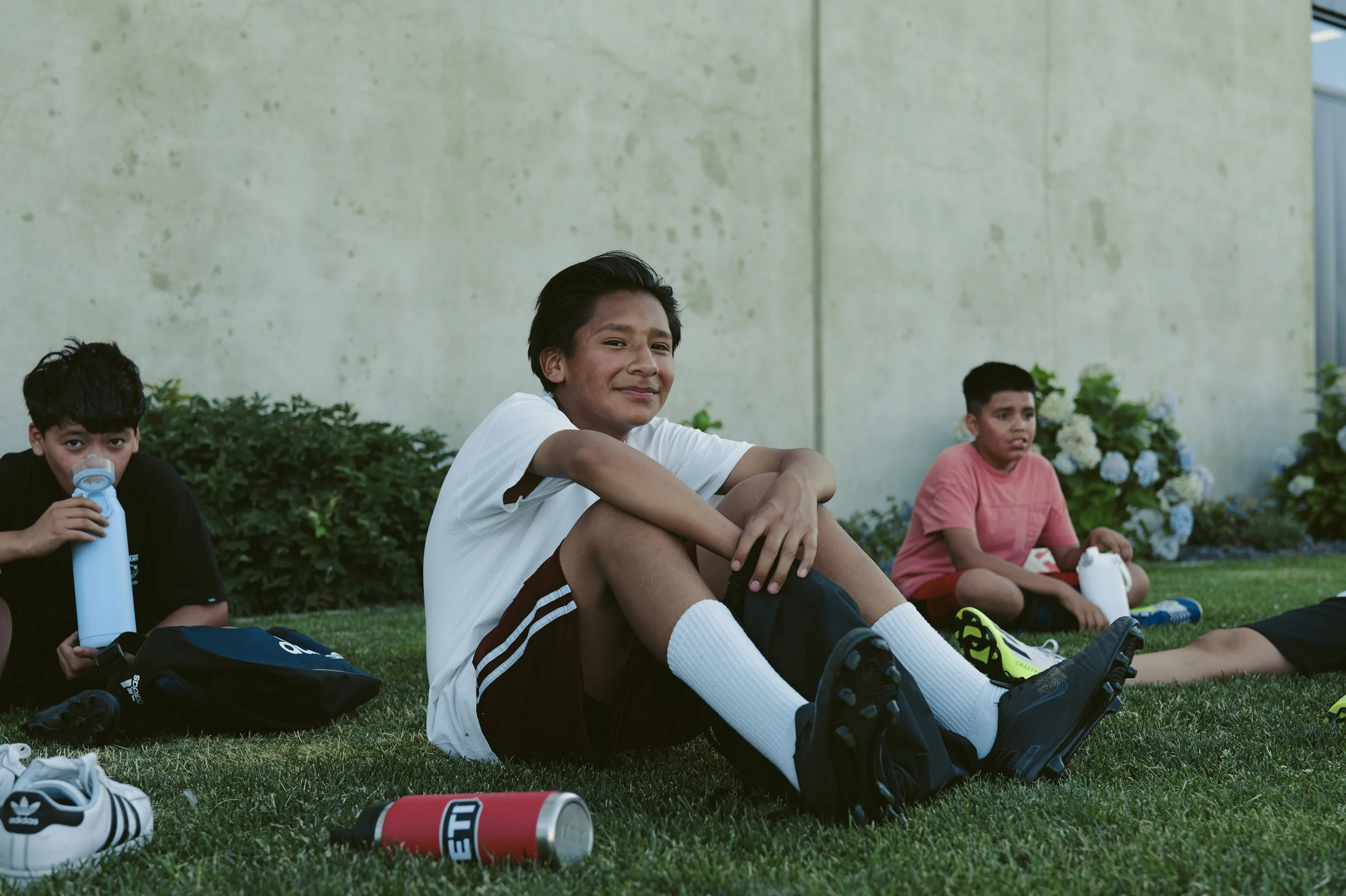 Three young boys sitting on grass outside near a beige wall on a sunny day, with water bottles and sports shoes around, some of them smiling and others looking serious.