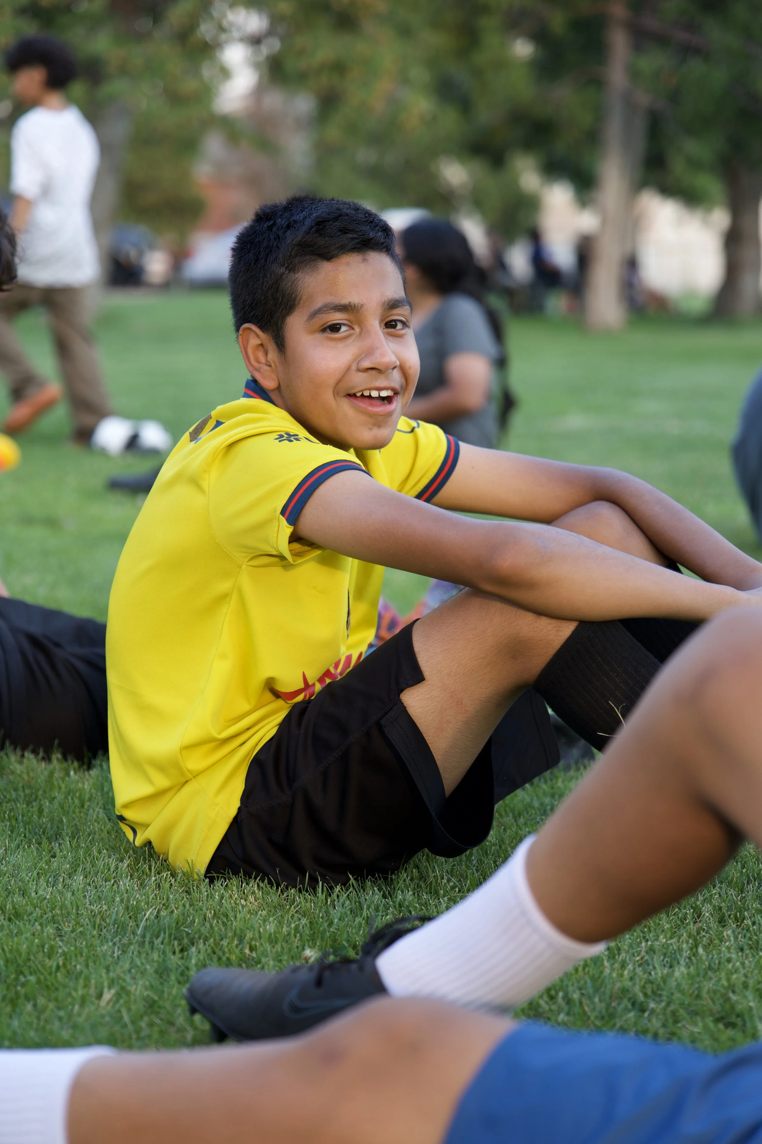 A young boy in a yellow sports jersey and black shorts sitting on the grass at a park, smiling at the camera with other people and trees in the background.
