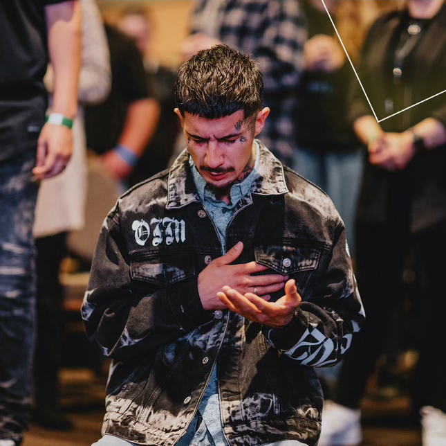 A young man with tattoos on his face and hand, wearing a black denim jacket, is standing with his eyes closed and one hand on his chest, while his other hand is raised with an open palm. He appears to be in a moment of prayer or reflection, surrounde