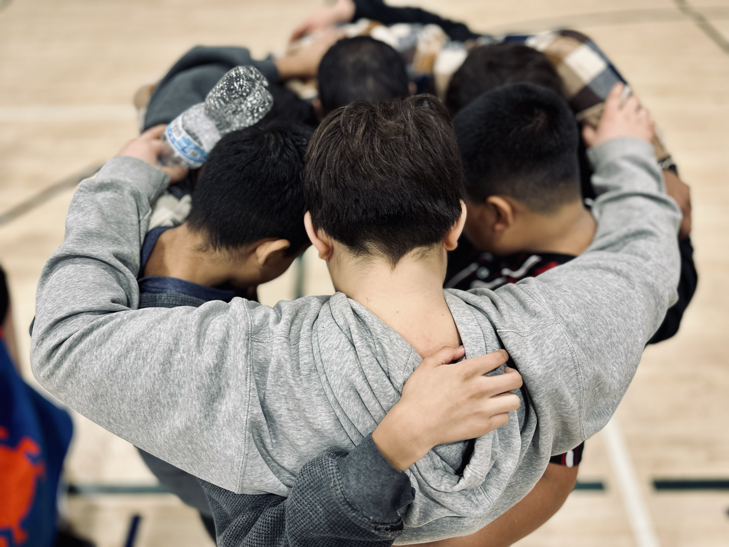 A group of boys in a circle with their heads down, embracing each other in a huddle on a gymnasium floor.