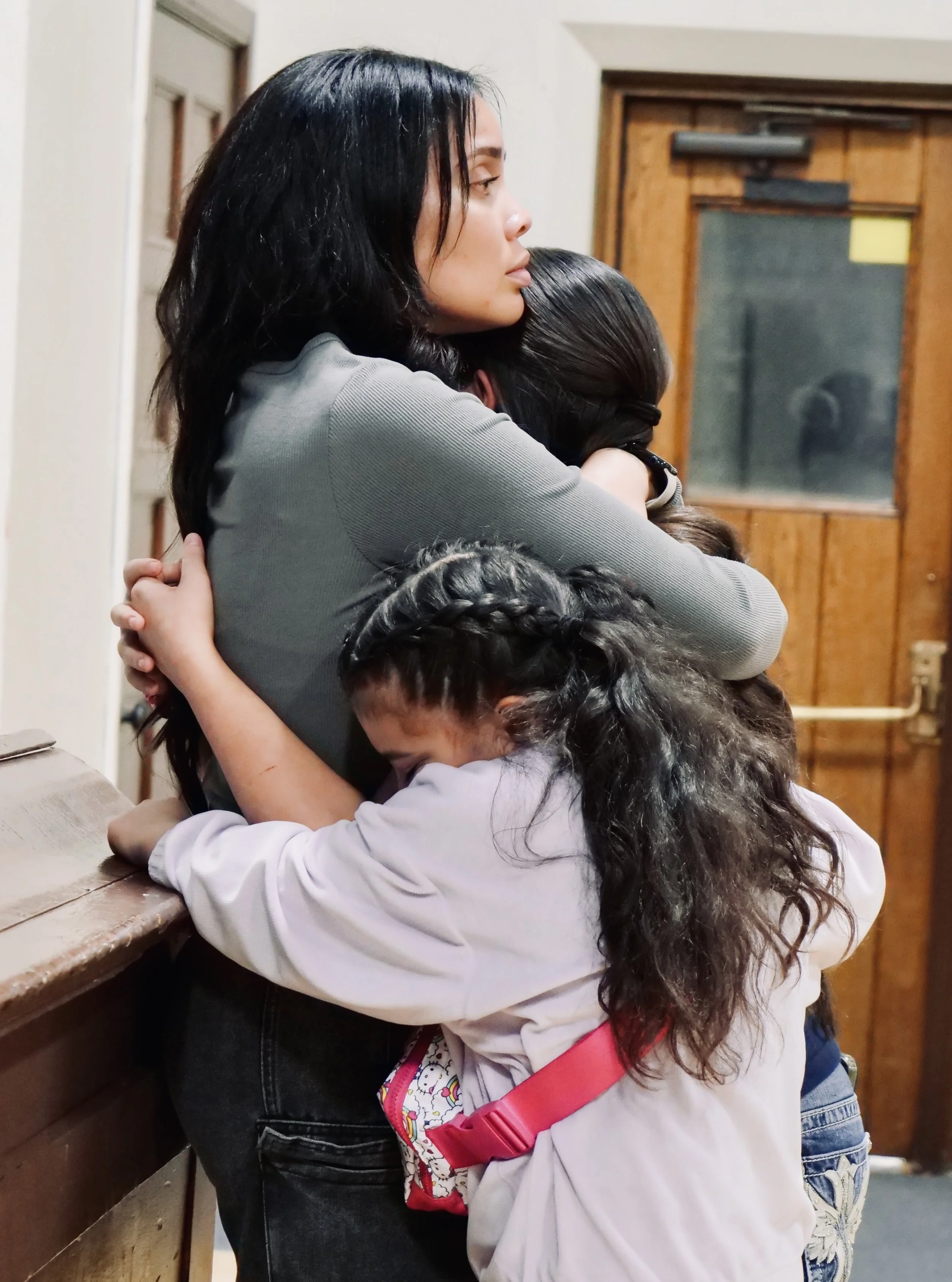 A woman with long black hair hugging two young girls, one with braided hair and the other with curly hair, inside a room with wooden doors and a window in the background.