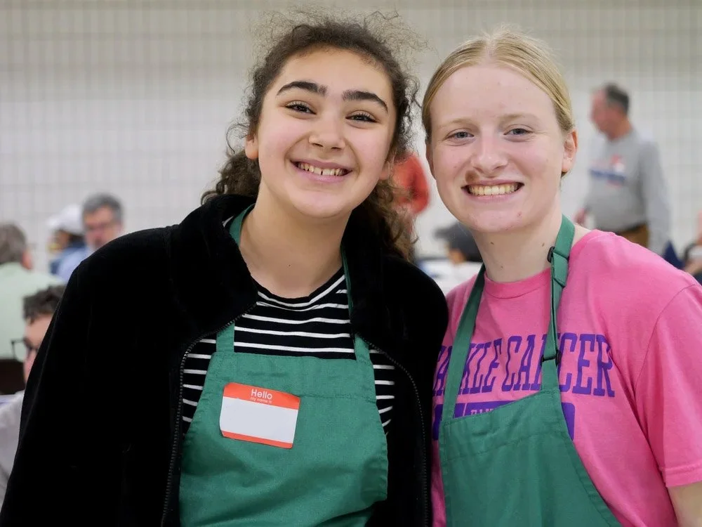 Two young women smiling, wearing green aprons, in a crowded indoor setting.