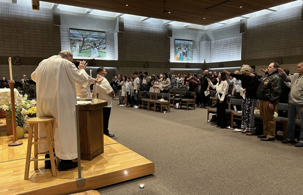 A church congregation is participating in a communion service, with the priest and altar servers at the front and the congregation standing with right arms extended.